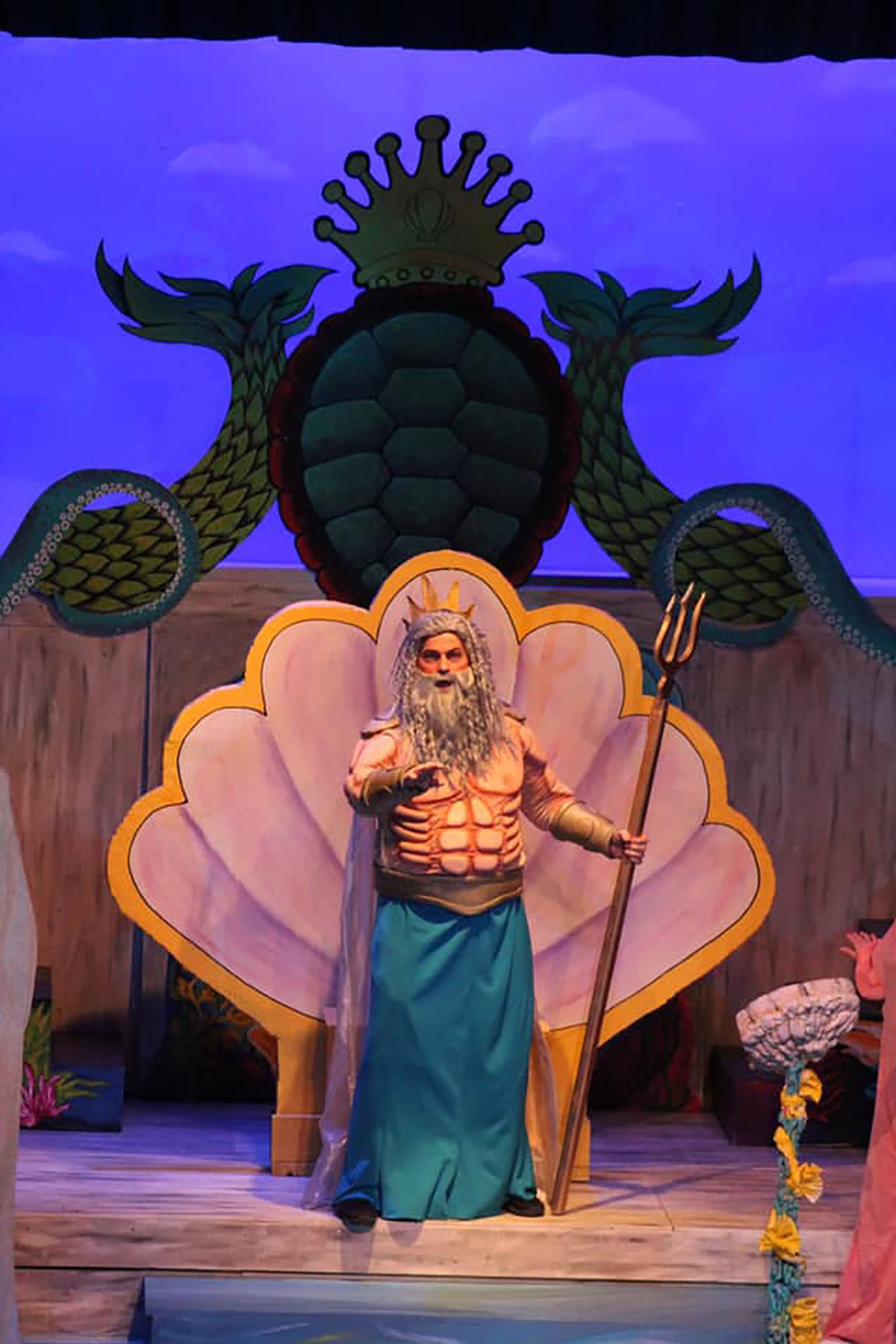 Performer dressed as Poseidon on stage with ocean-themed backdrop and props.
