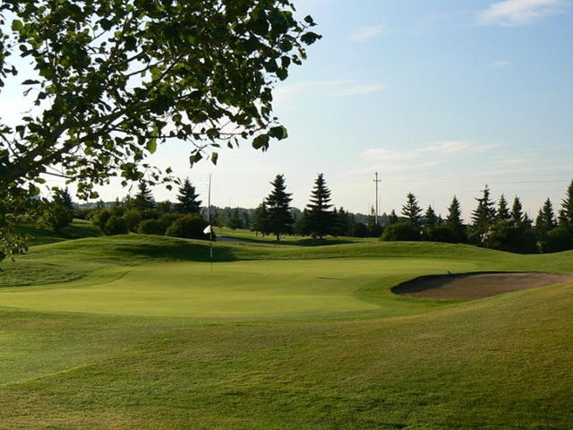 Golf green at Fox Hollow with trees and a sand bunker under a clear sky.