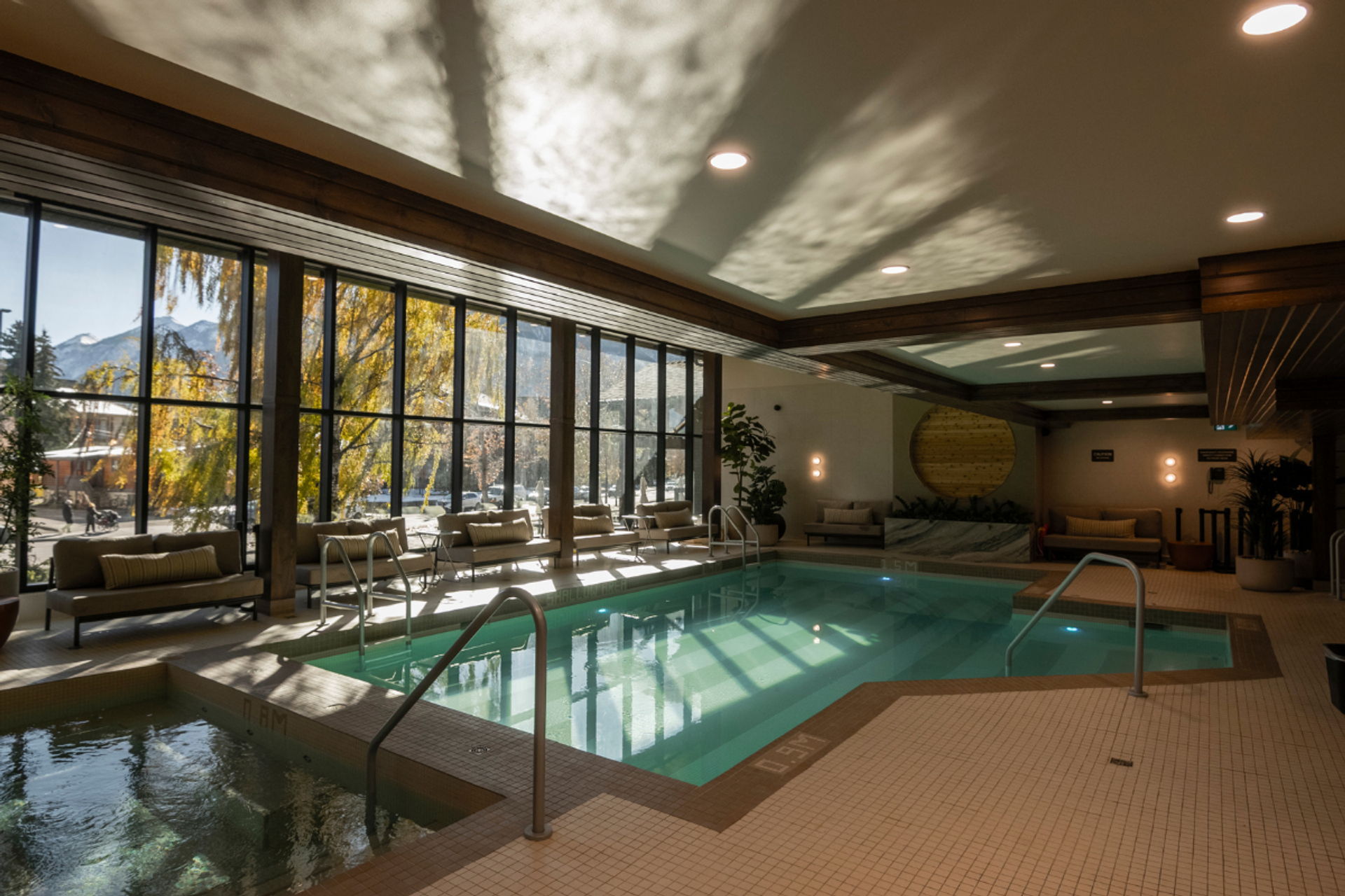 Indoor swimming pool and hot tub with large windows revealing autumn trees and mountains, surrounded by lounge seating.