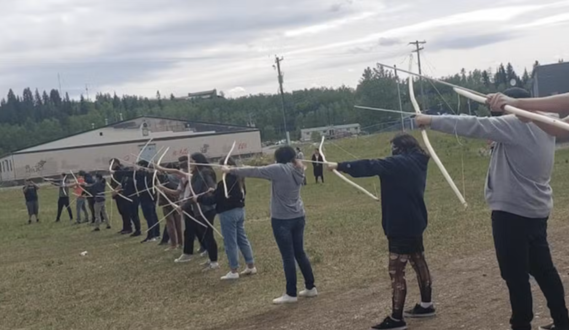 Line of people on grassy field aiming white bows toward unseen archery targets.