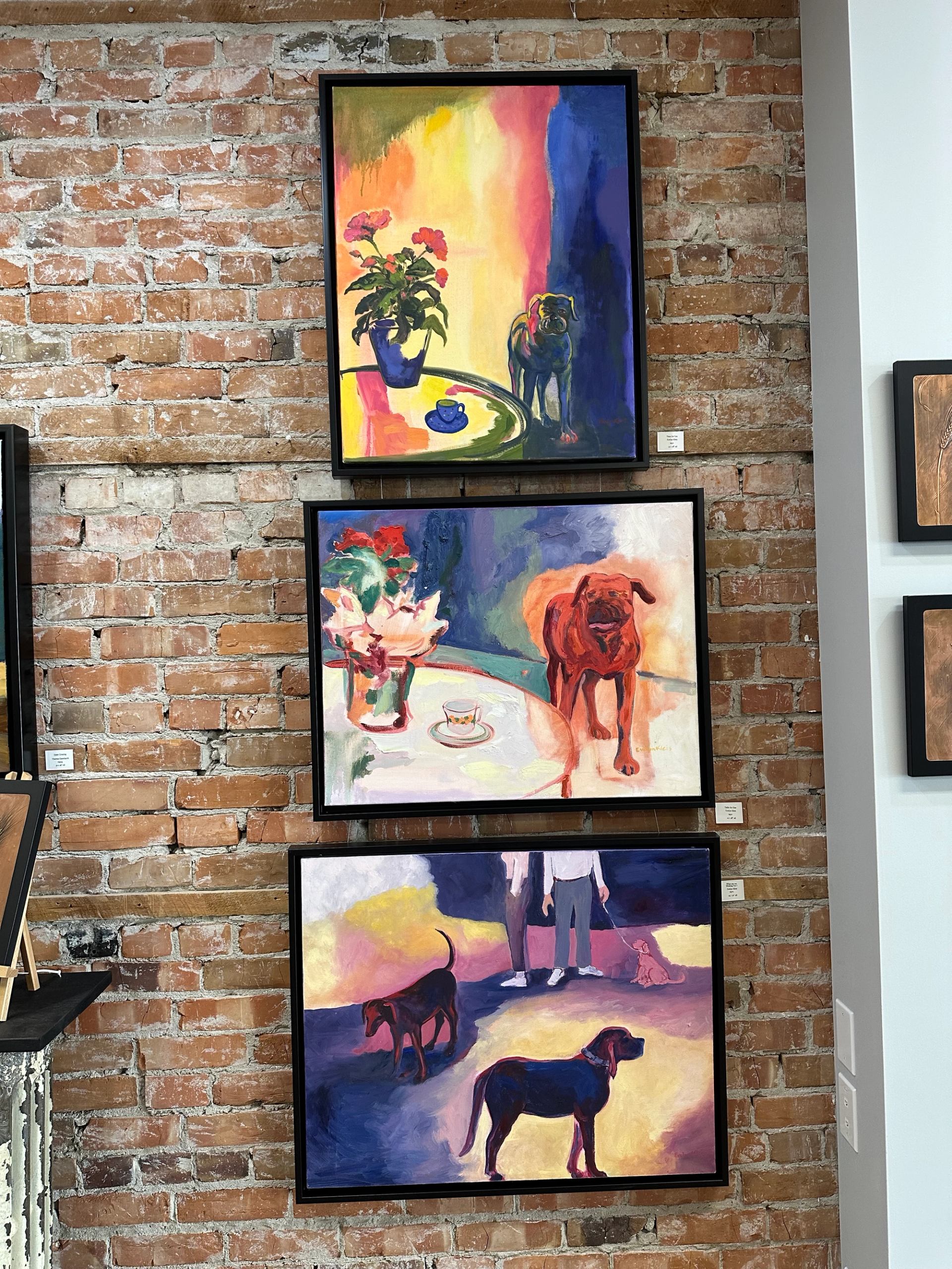 Colorful paintings of dogs and still life scenes hung vertically on a brick wall.