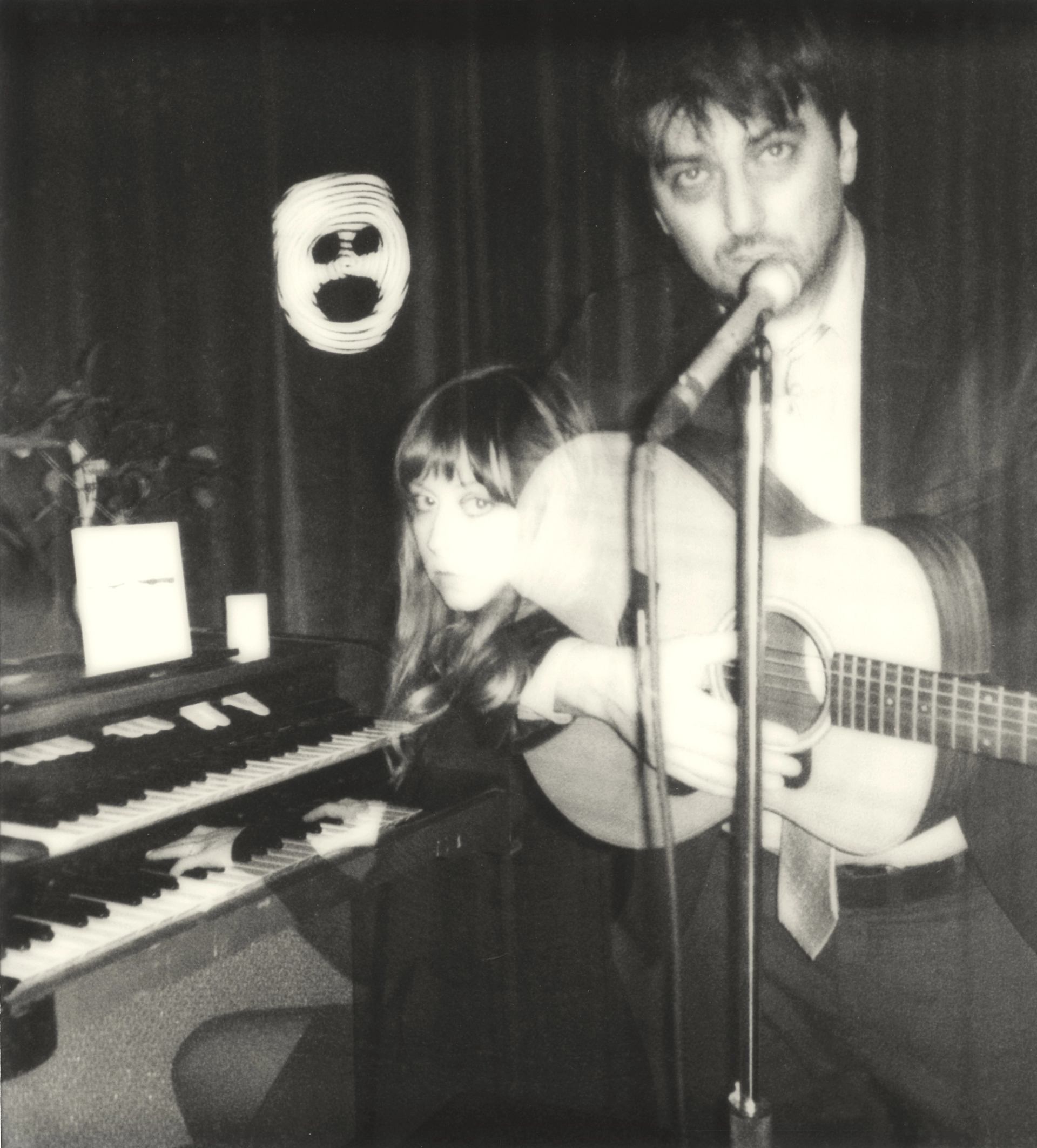 Two musicians perform indoors—one on guitar and vocals, the other on keyboard, with a mask on the wall.