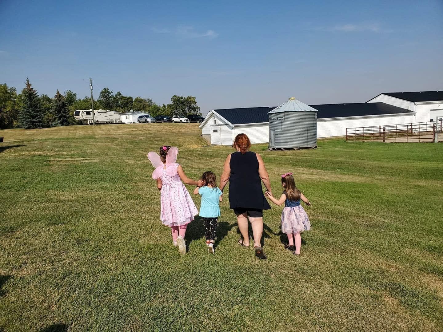 An adult walks across a grassy field holding hands with three children near farm buildings.