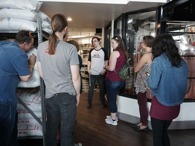 Tour group stands in brewery tasting room around stainless steel brewing equipment and barrels.