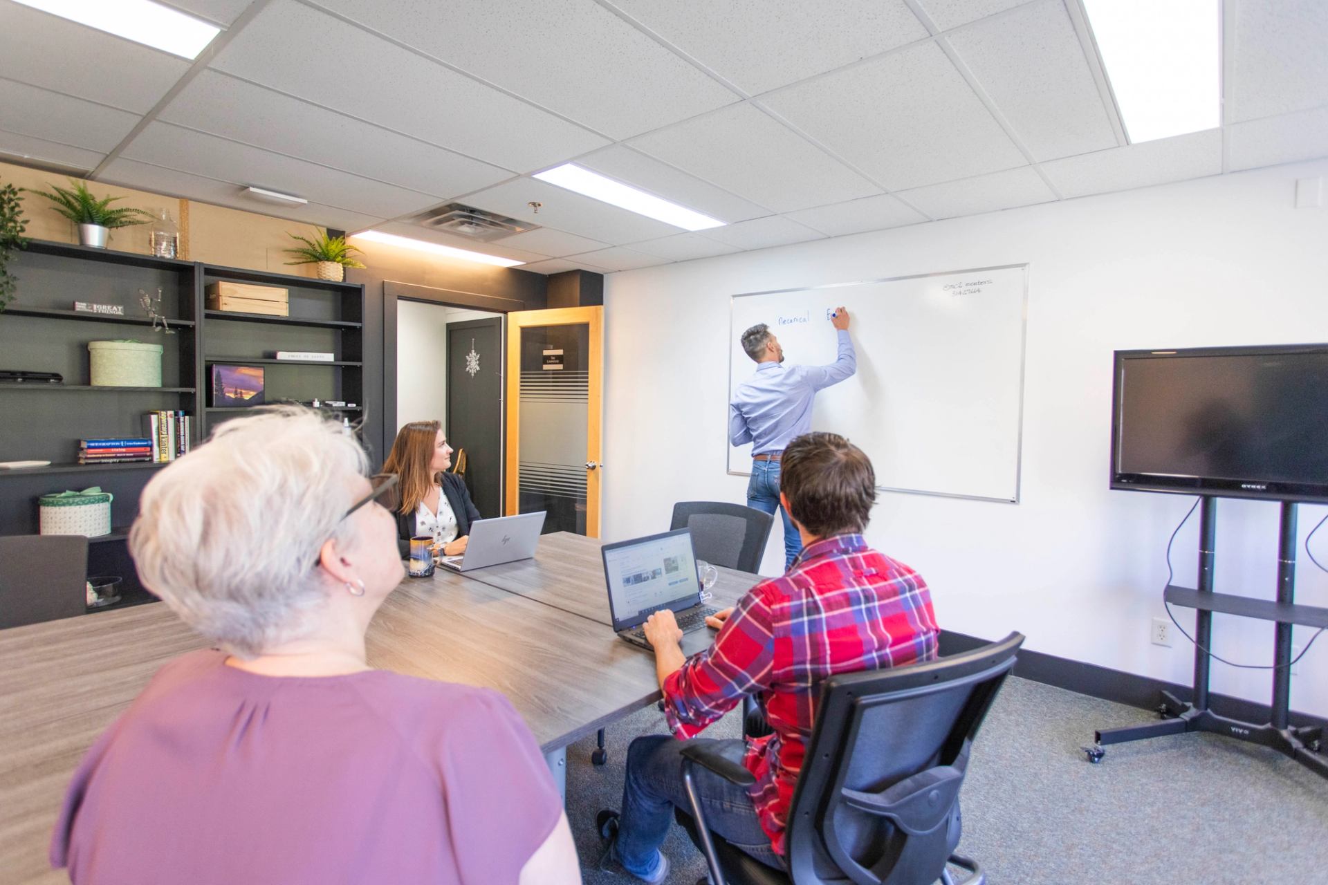 Four people in a meeting. One stands at large whiteboard, in front others sitting at a table.