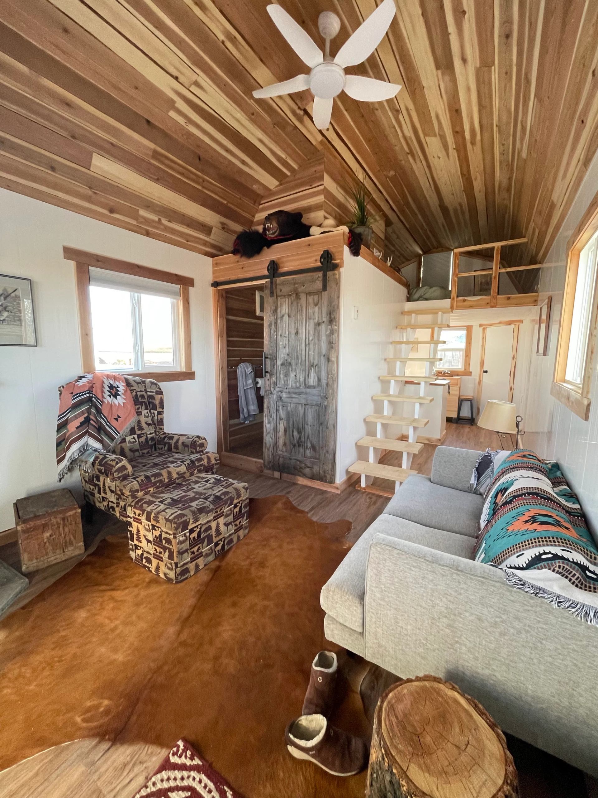 Interior of a cabin with wood-paneled ceiling, cozy seating, and a loft above.