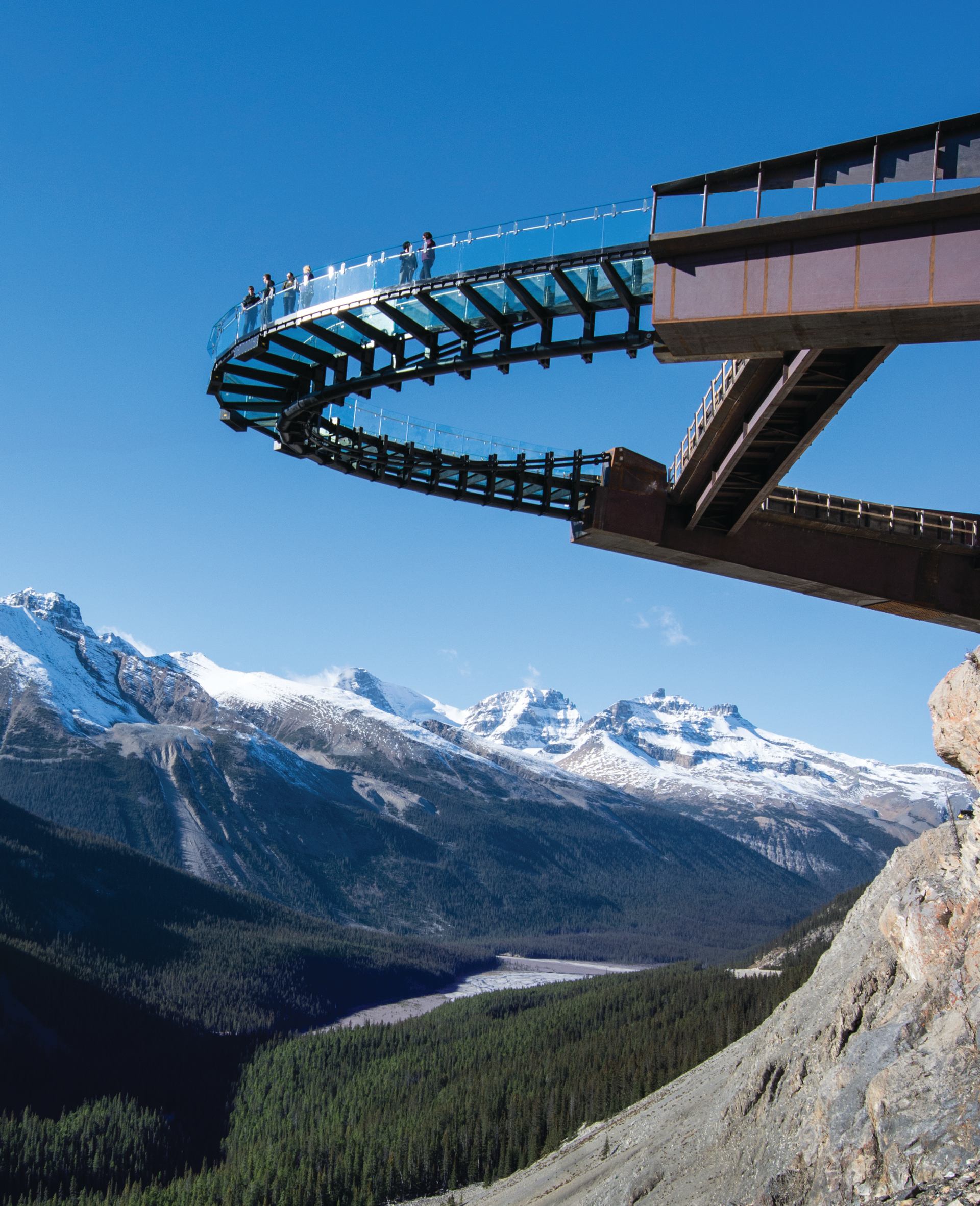 Glass-floored cliffside walkway curves over forested valley with distant snow-covered mountains.