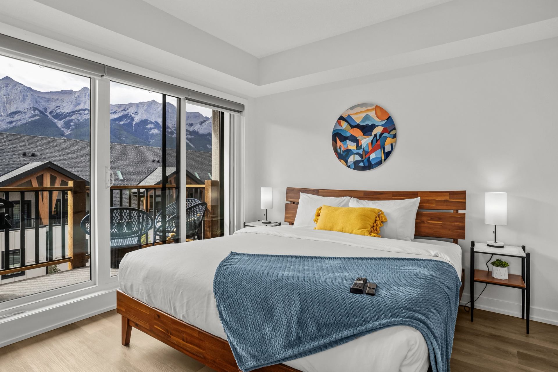Bedroom with large window, mountain view, wooden bed, blue blanket and balcony chairs.