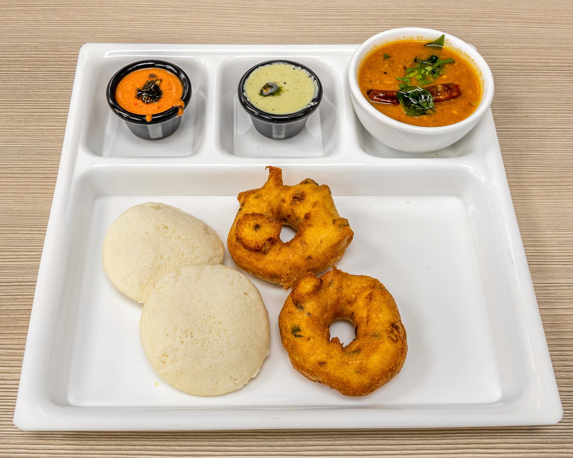 Idli and vada served with sambar and two chutneys on a white tray.