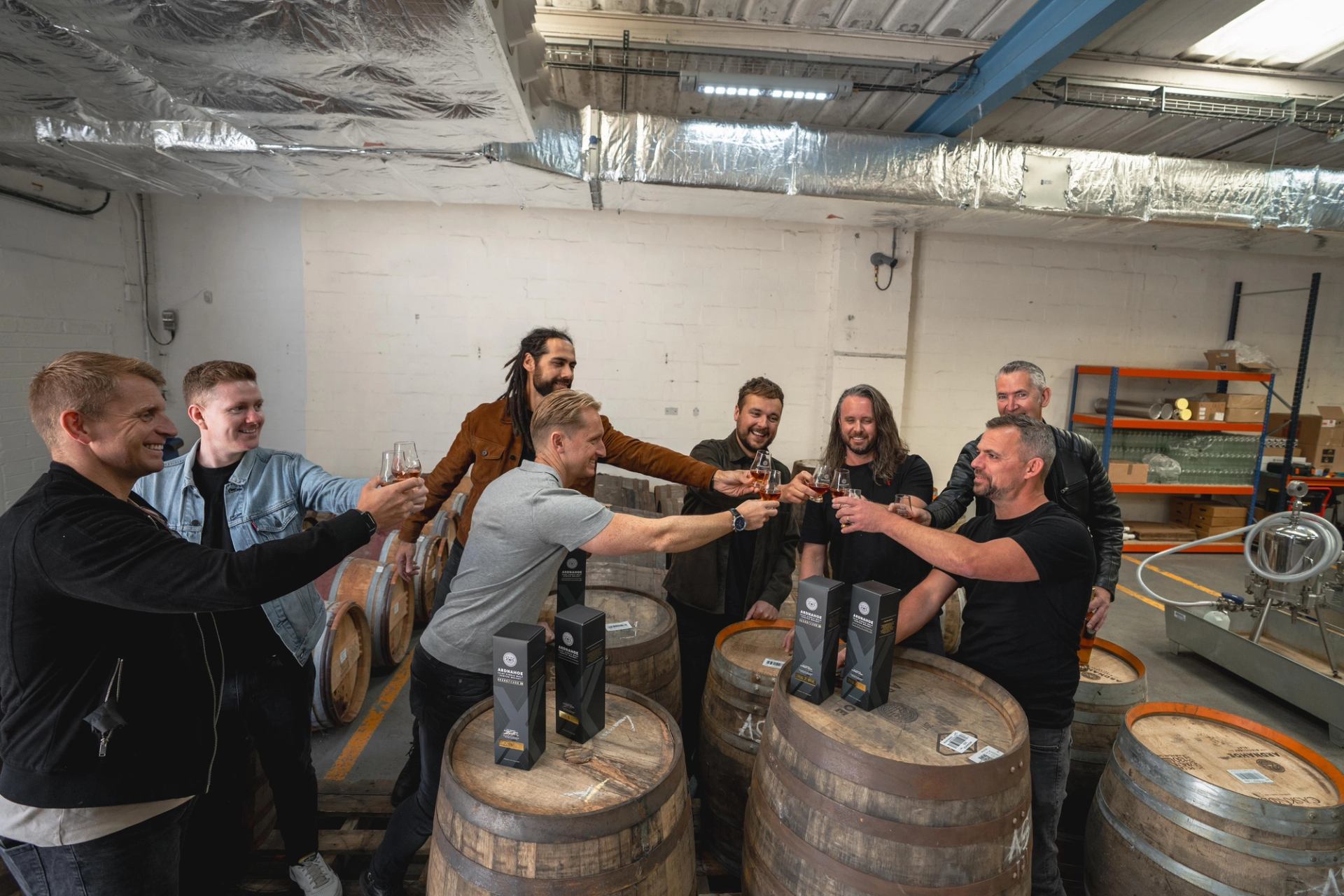 Group of people toasting with whisky glasses around wooden barrels in a distillery.