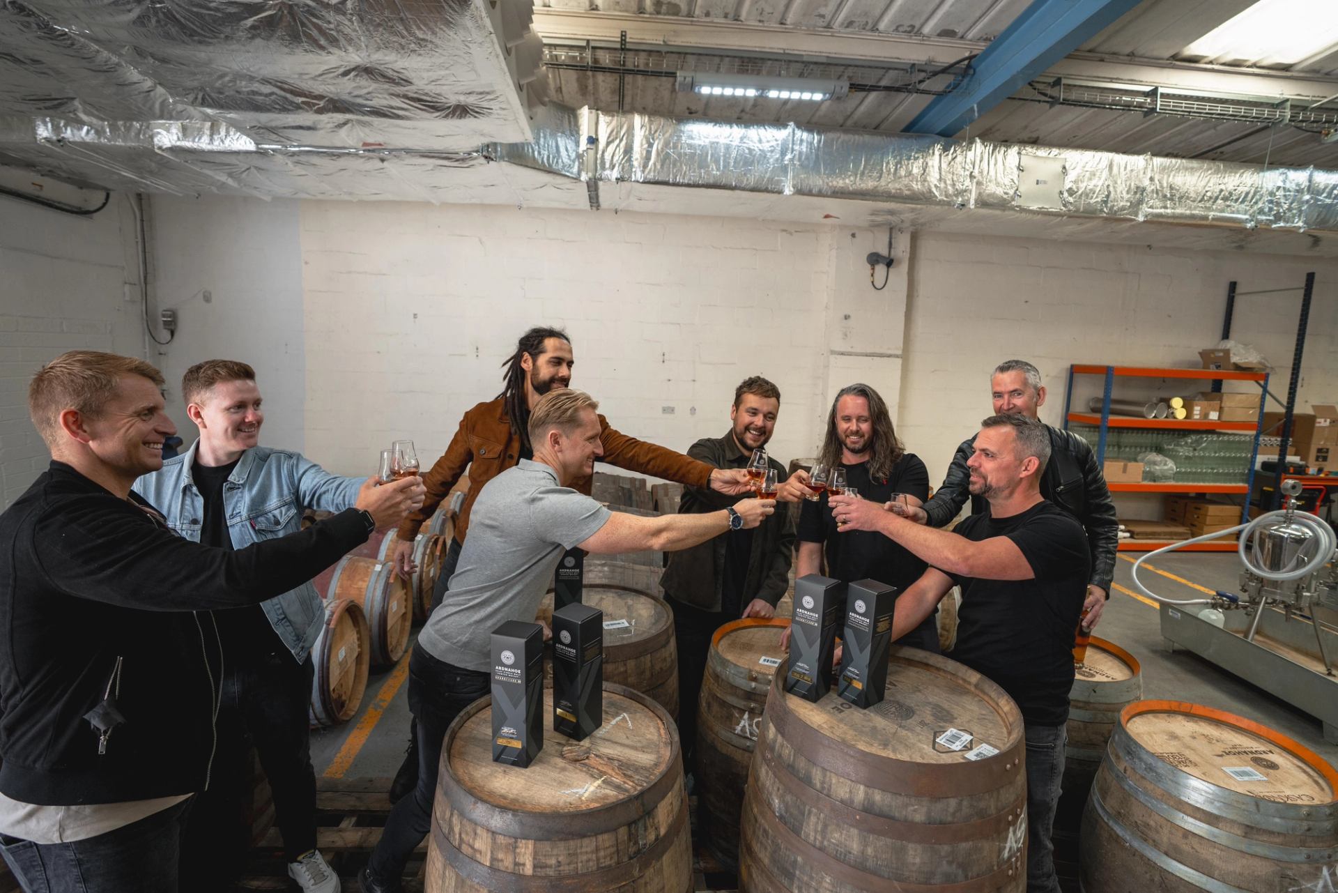 Group of people toasting with whisky glasses around wooden barrels in a distillery.