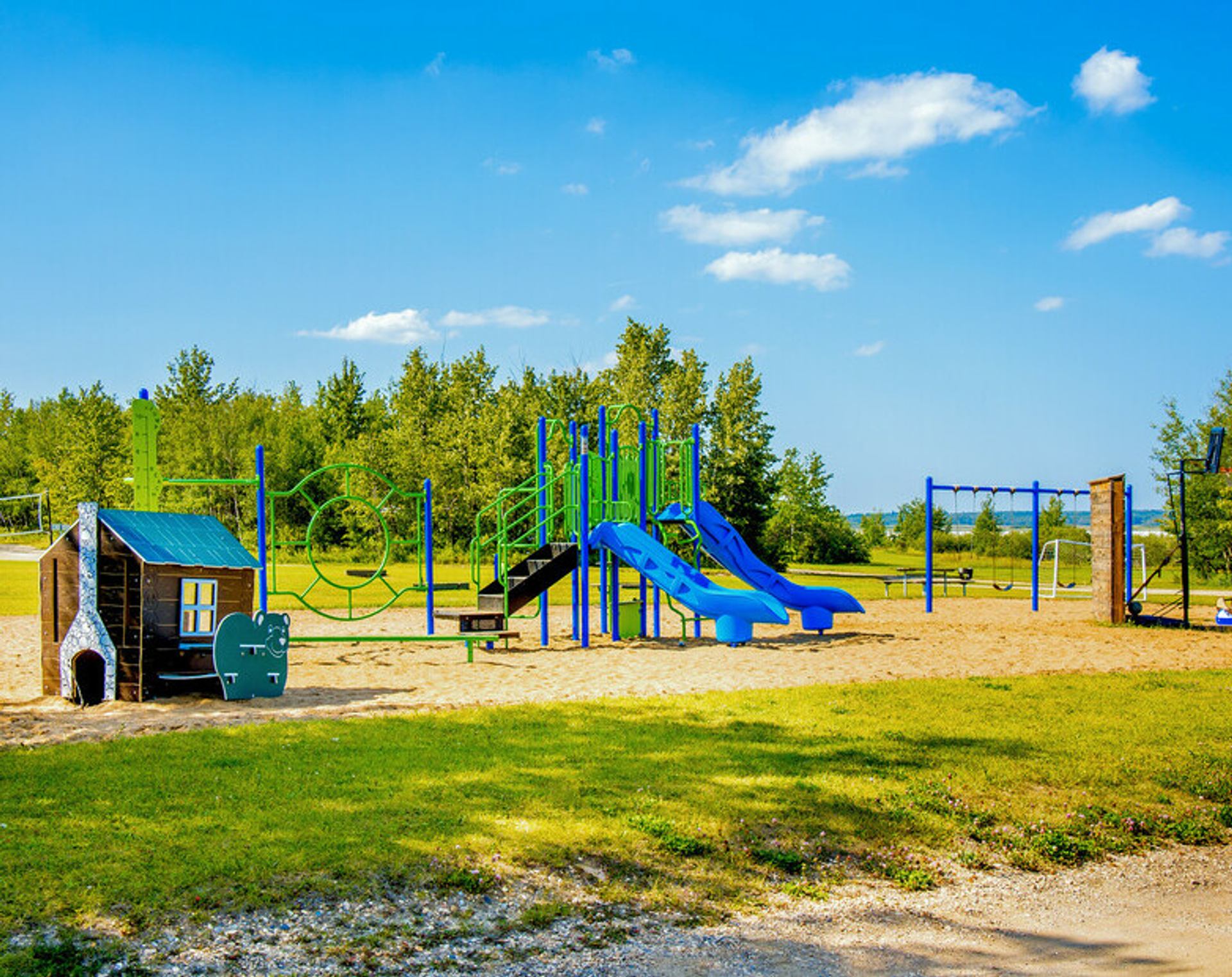 A sunny playground with a wooden playhouse, blue slides, and swings on sandy ground.