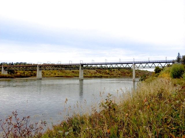 A long pedestrian bridge stretches across a calm river beside grassy banks.