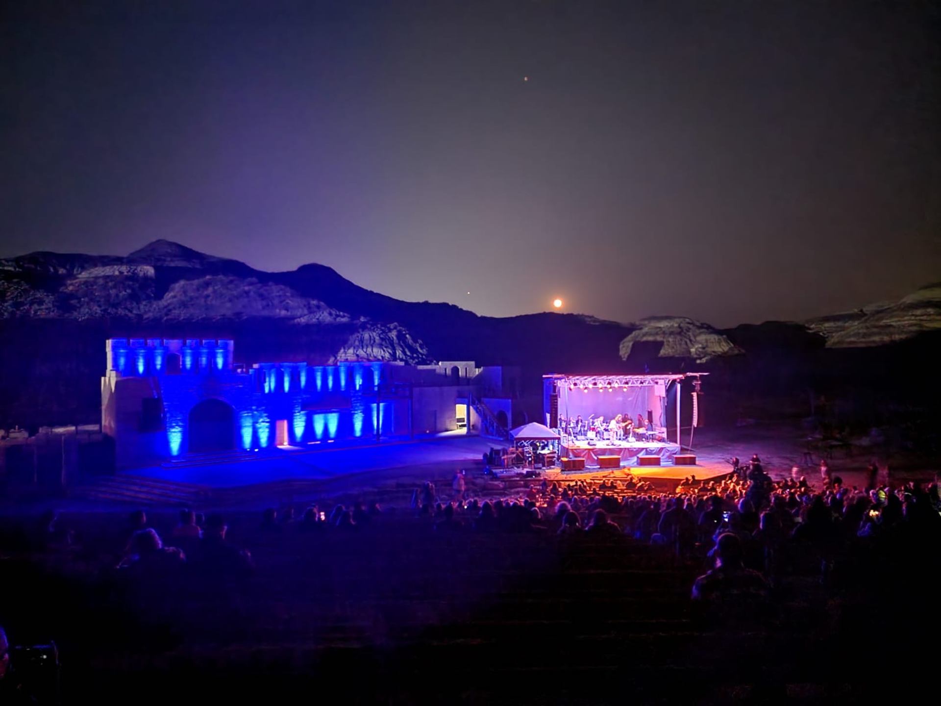 Night concert with stage lights, audience, and mountain backdrop.