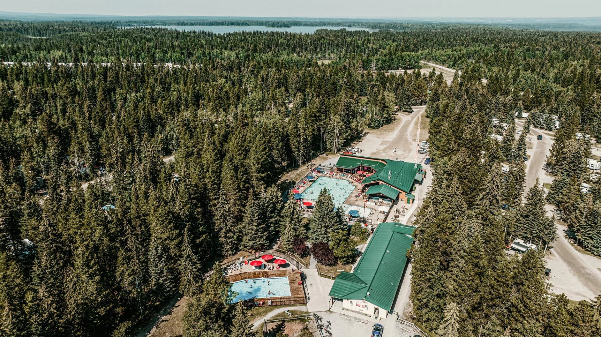 Aerial view of forested campground with outdoor swimming pools, green-roofed buildings and nearby lake.