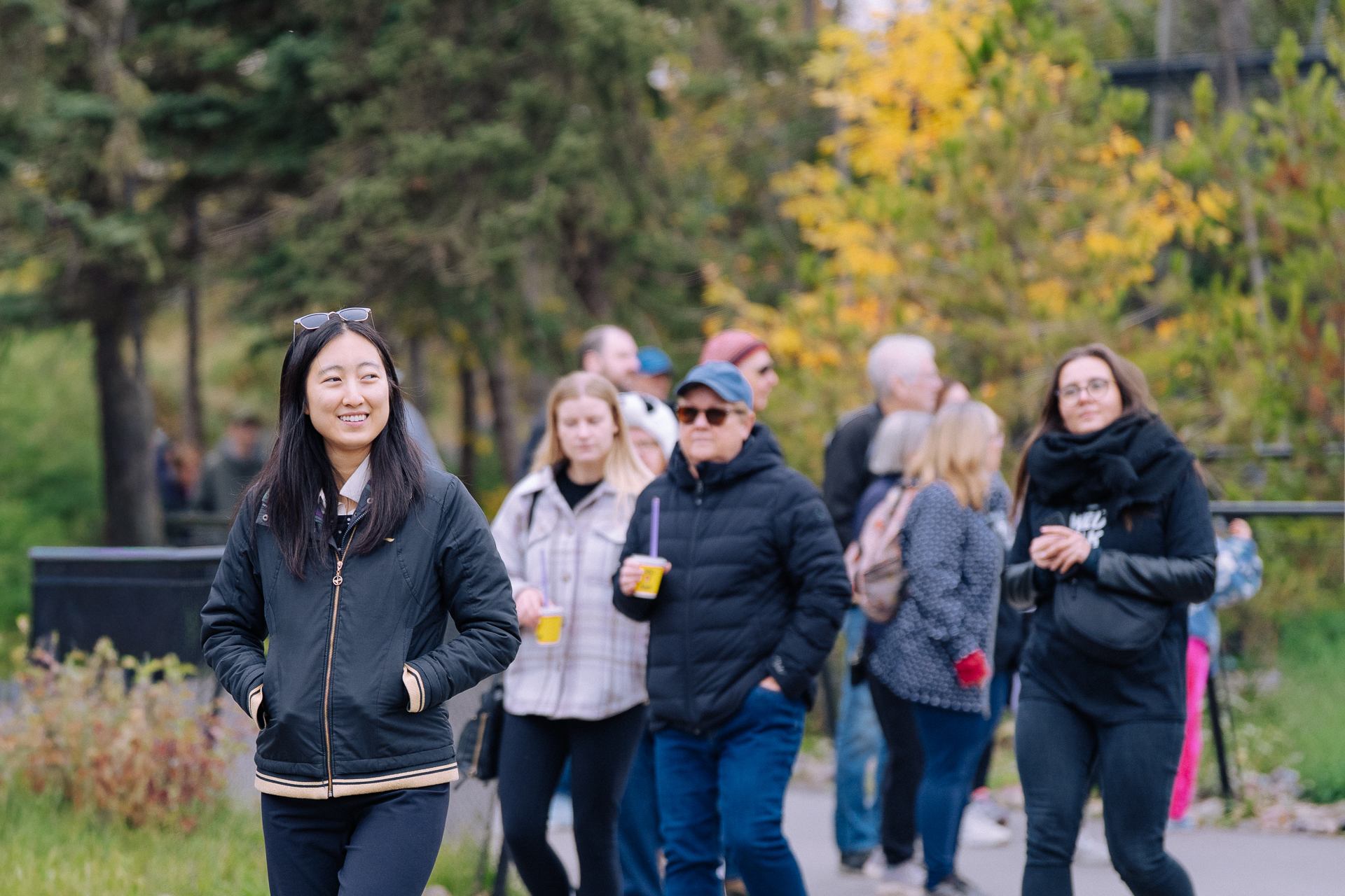 Several people walk along an outdoor pathway bordered by trees and fall foliage.