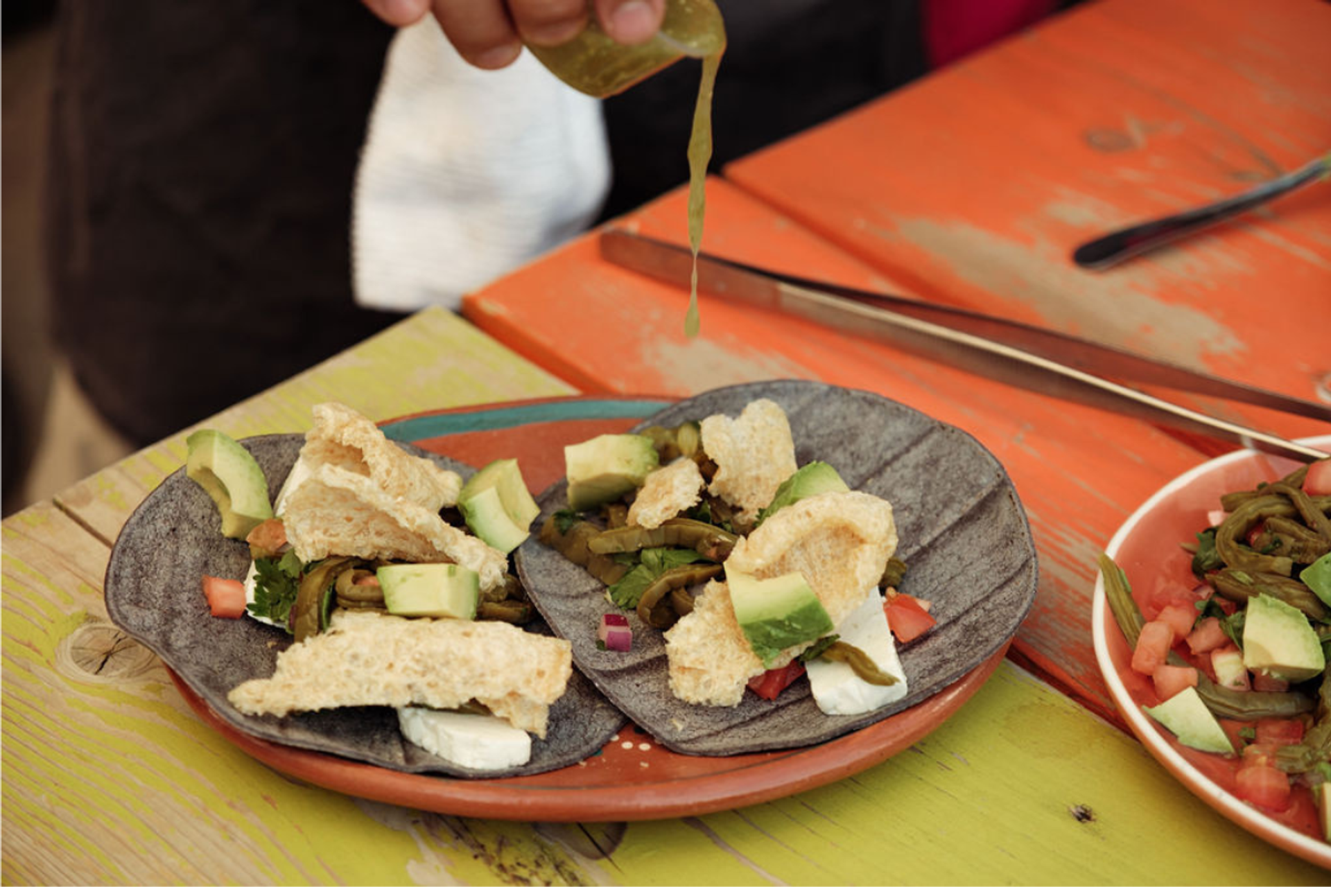 Blue corn tacos with avocado, grilled veggies, and green sauce on colorful table.