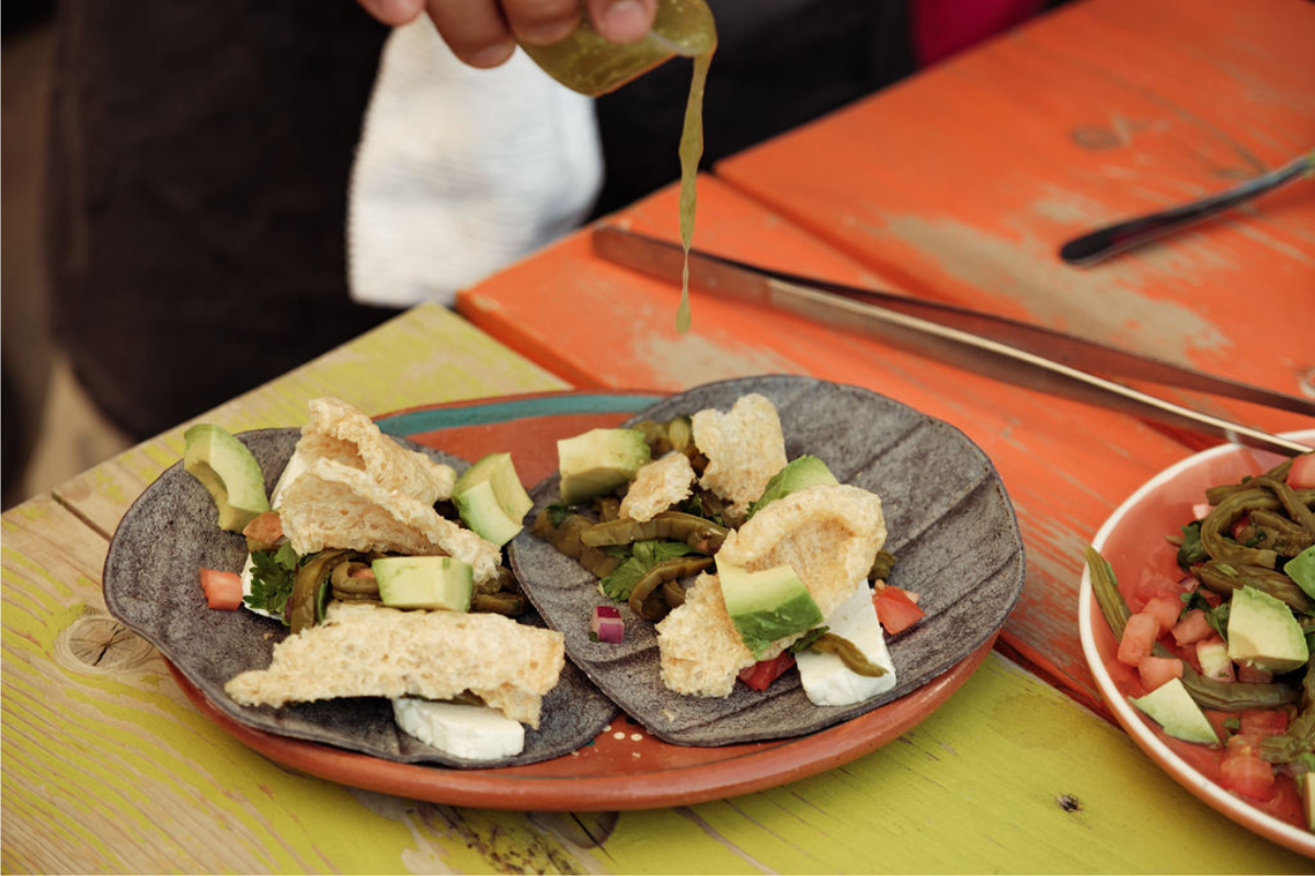 Blue corn tacos with avocado, grilled veggies, and green sauce on colorful table.