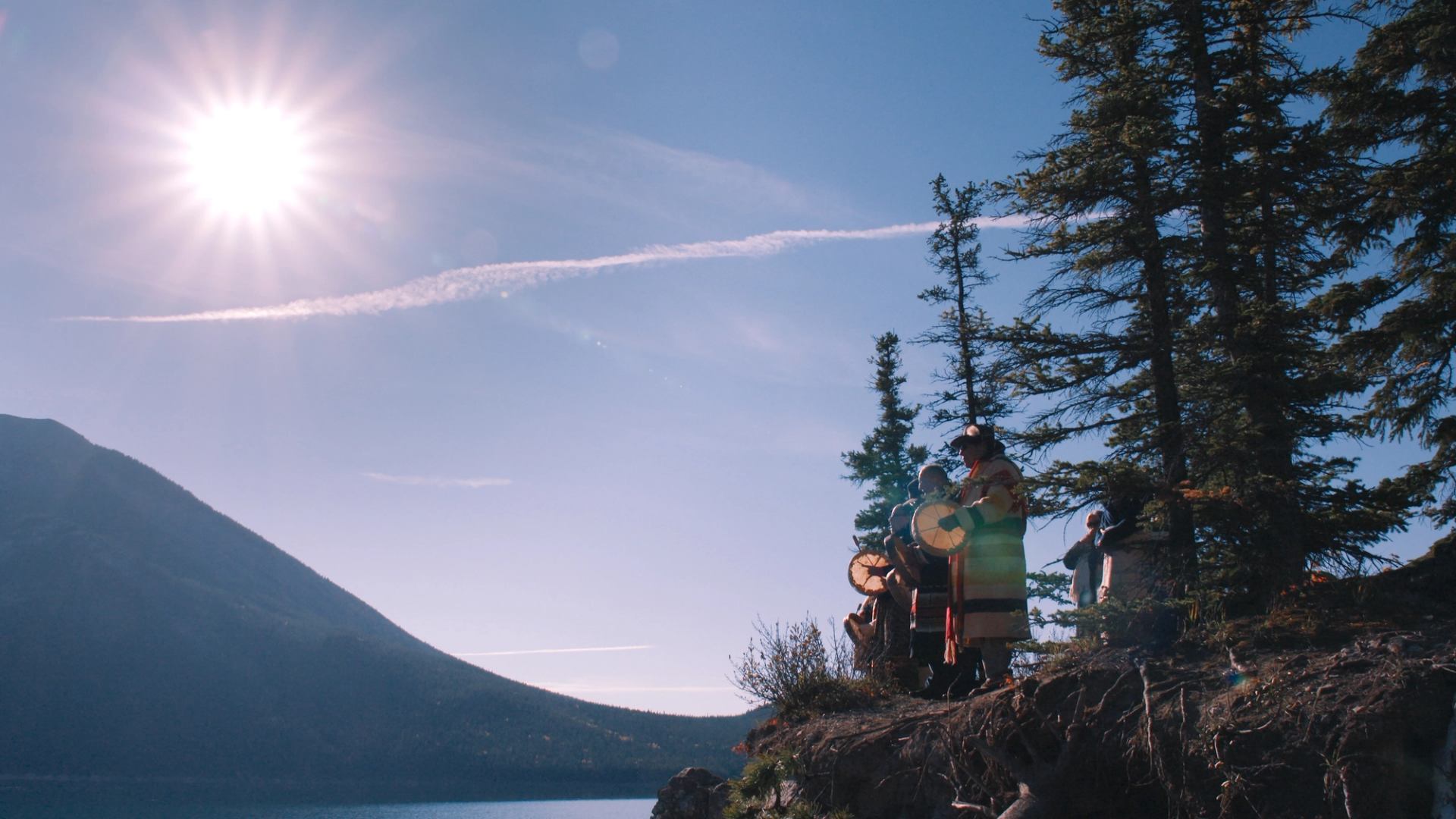 Group of people with hand drums stand on rocky lakeshore beside tall evergreens.