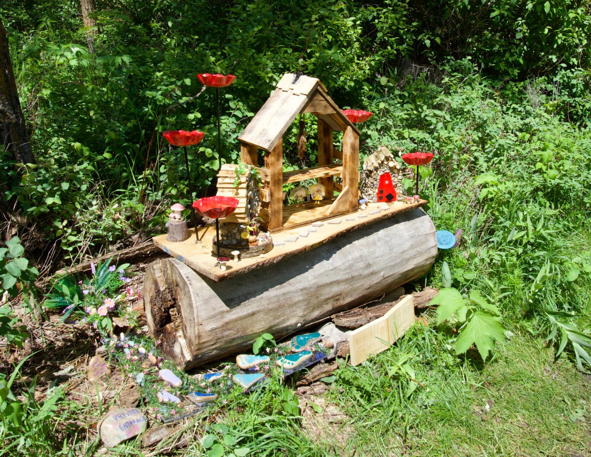 Wooden fairy house on fallen log with red glass flowers in green forest.