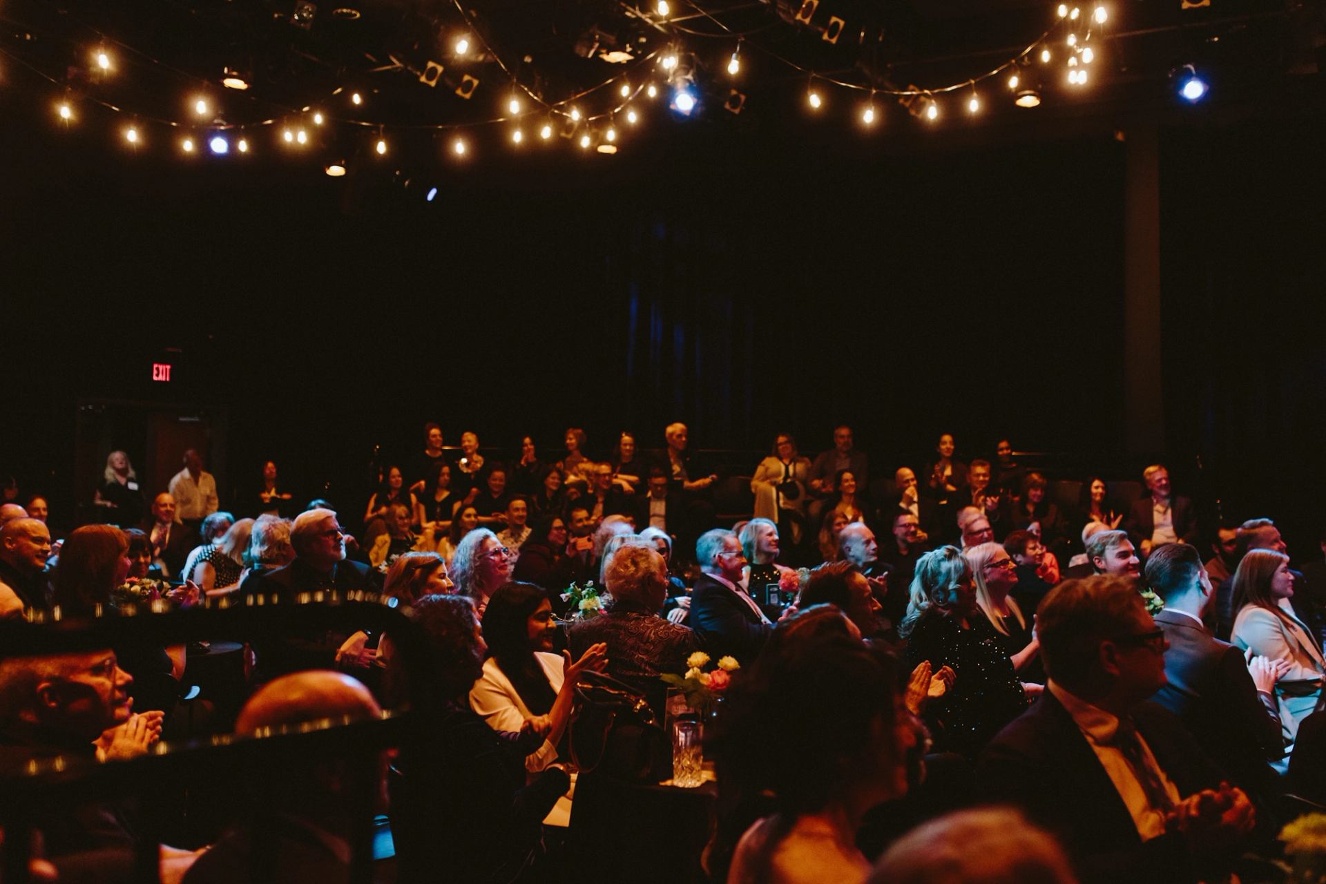 Audience seated in dim theatre, watching event under hanging string lights.