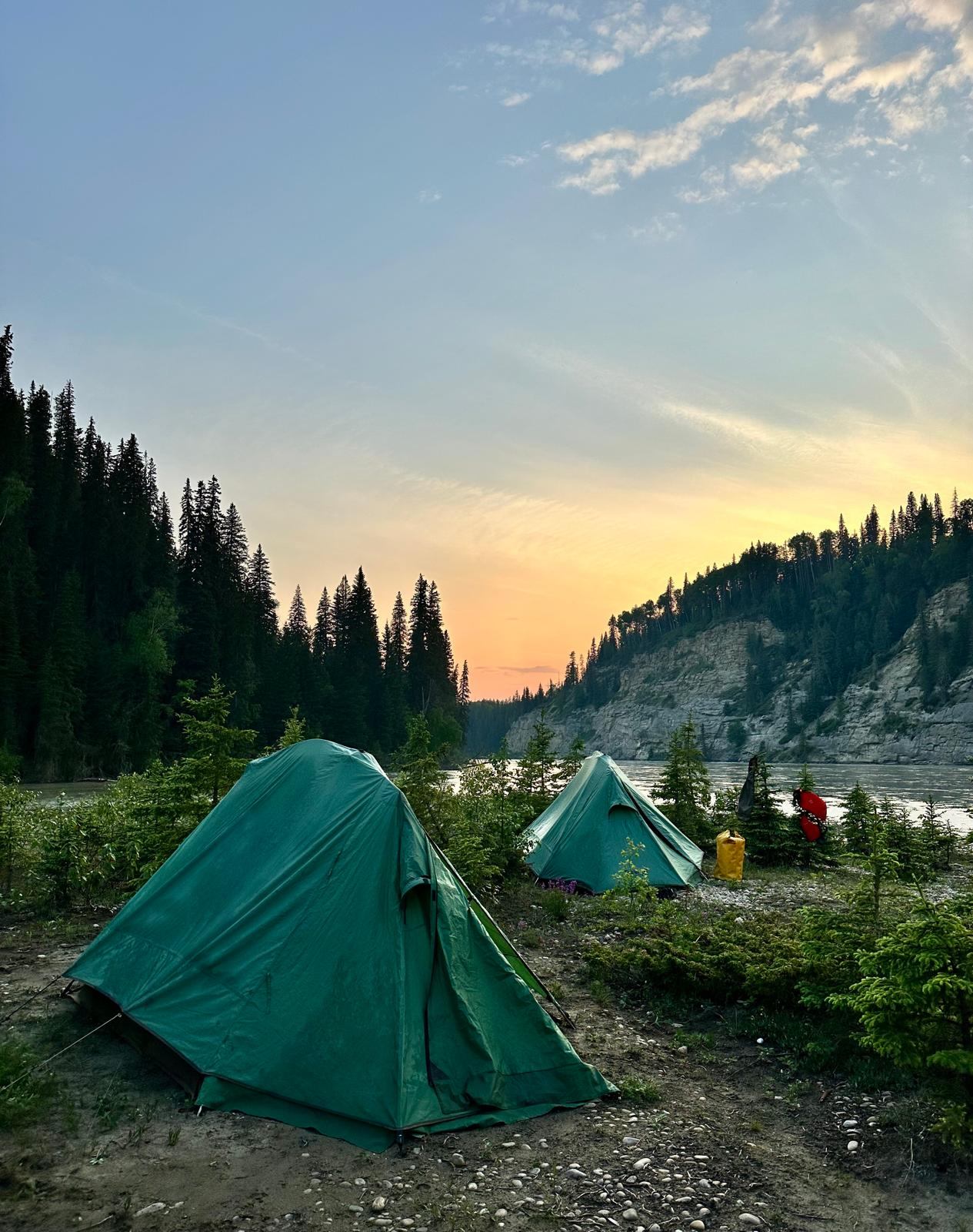 Sunsetting behind tall cliffs with green tents along the rocky shore of the Athabasca.