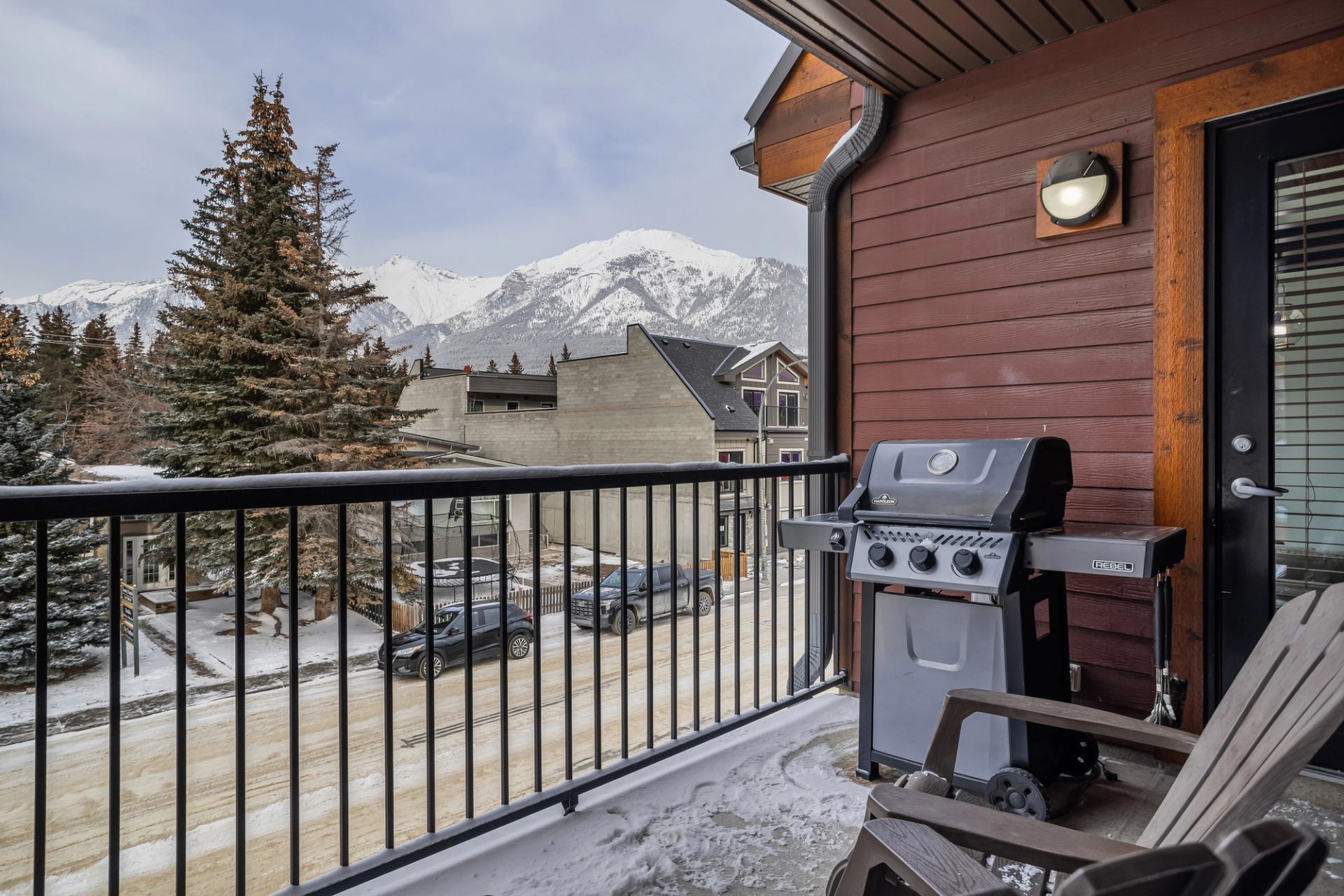 Balcony with barbecue grill overlooking snowy street, evergreen trees and mountain range.