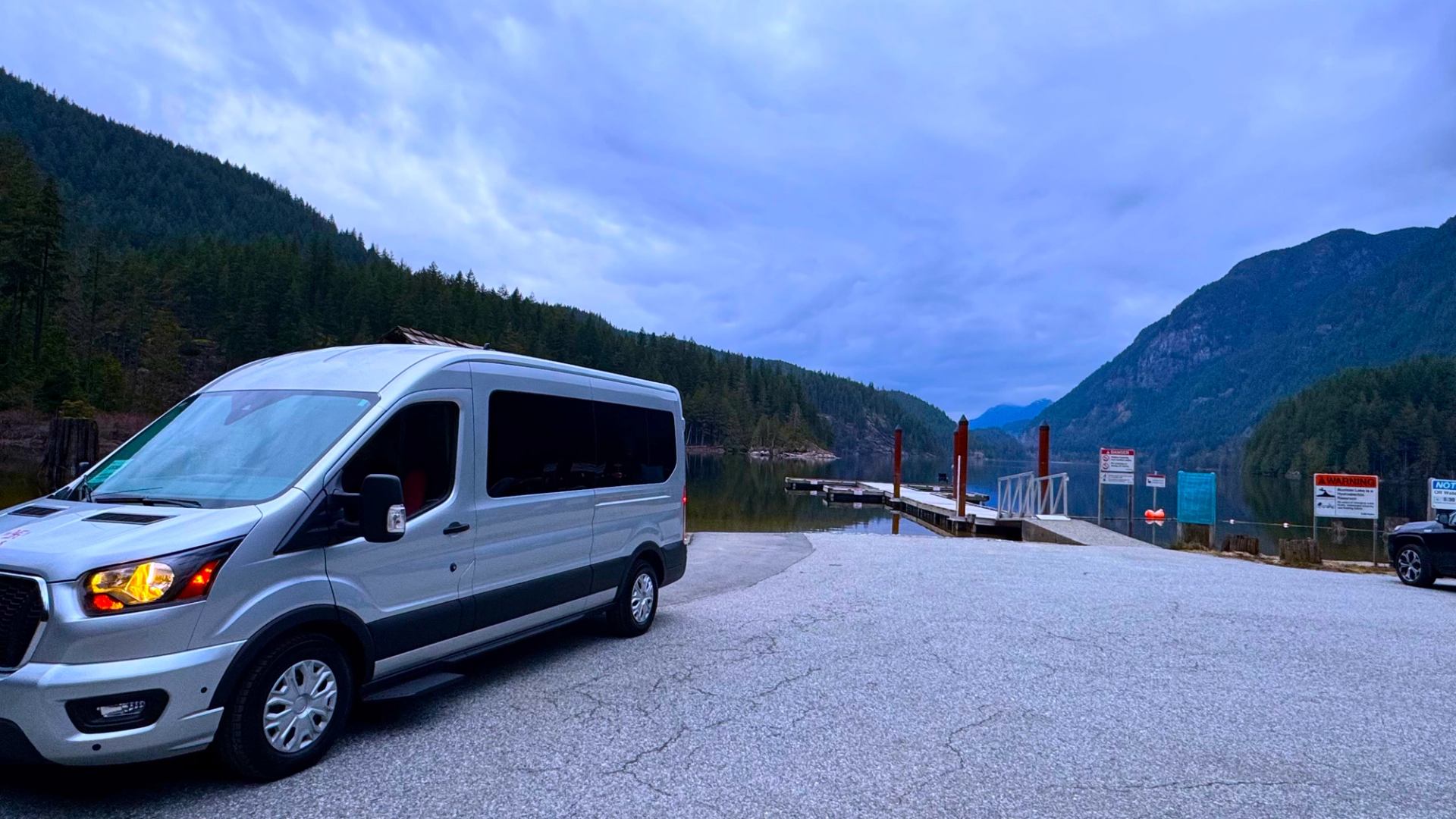 Silver passenger van parked near lakeside boat launch with forested mountains.