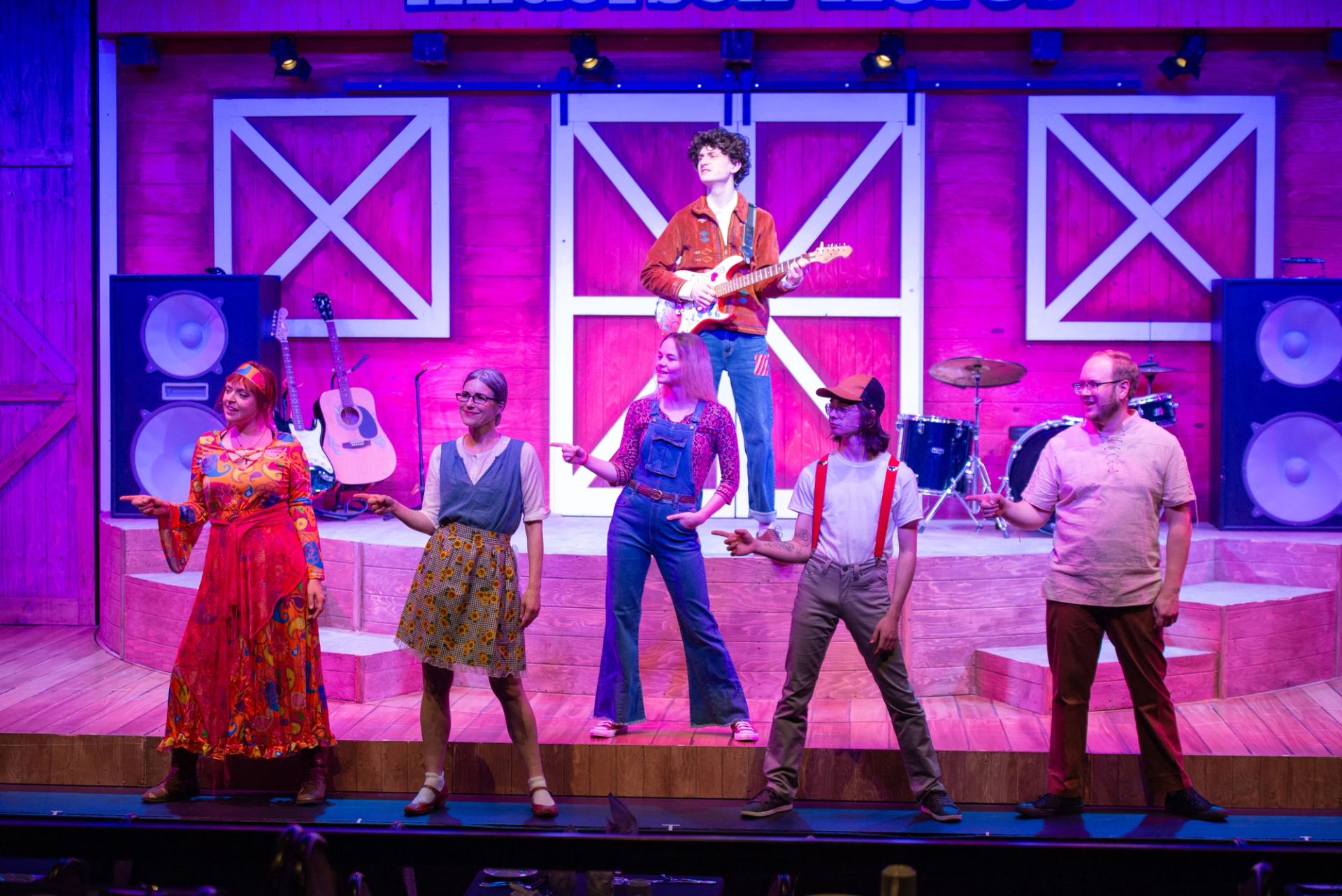 Stage performers in colourful costumes pose before barn-style set with guitars and drums.