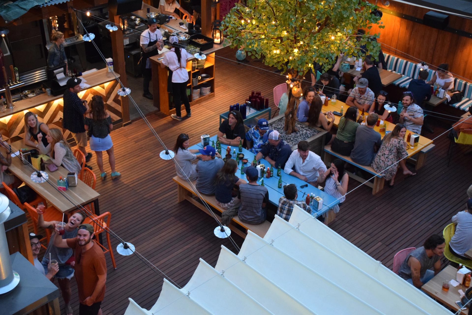 Lively rooftop bar scene with many people dining and socializing at tables, illuminated by string lights and a glowing tree.