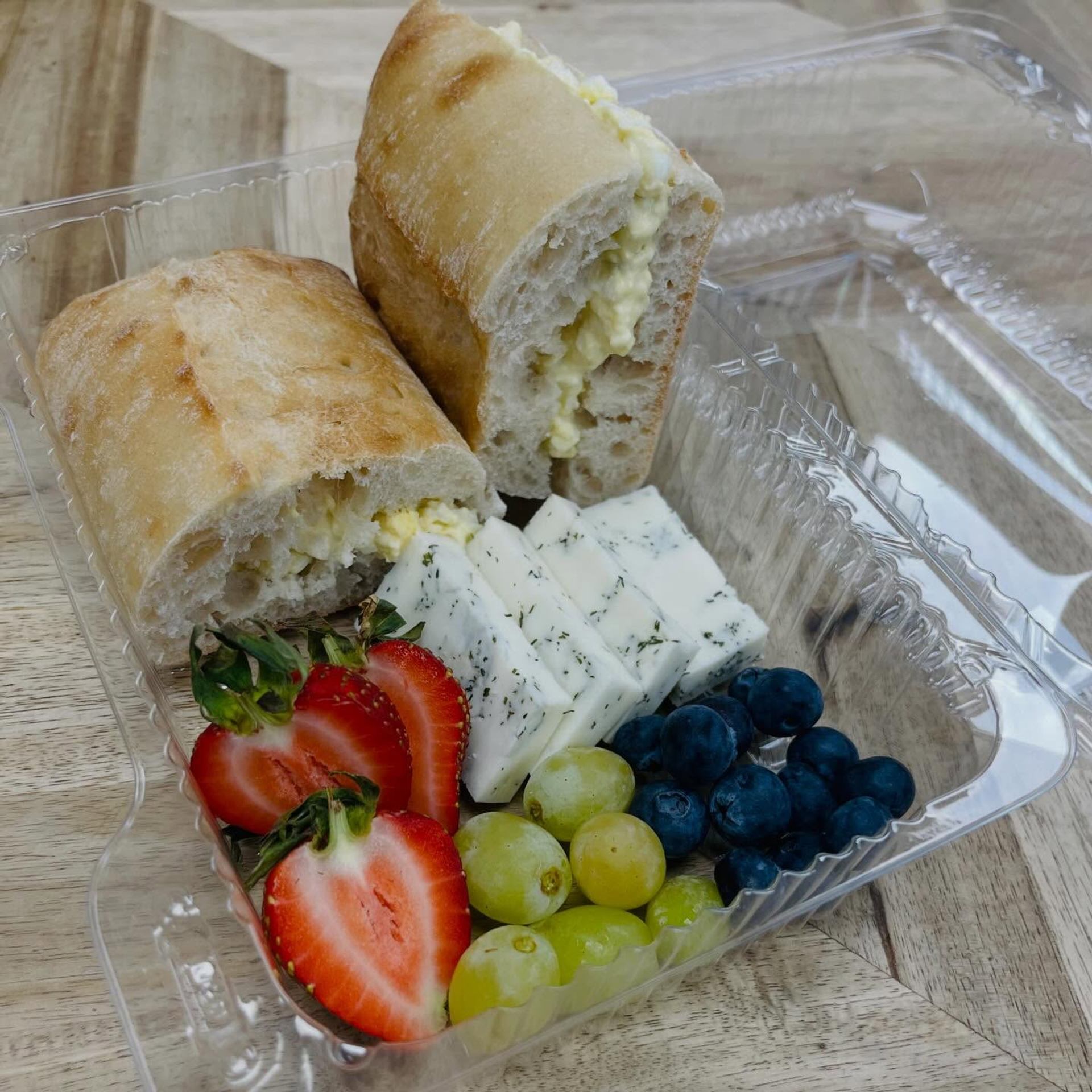 Fresh cheese, fruit, and a stuffed sandwich are arranged in a takeout container.