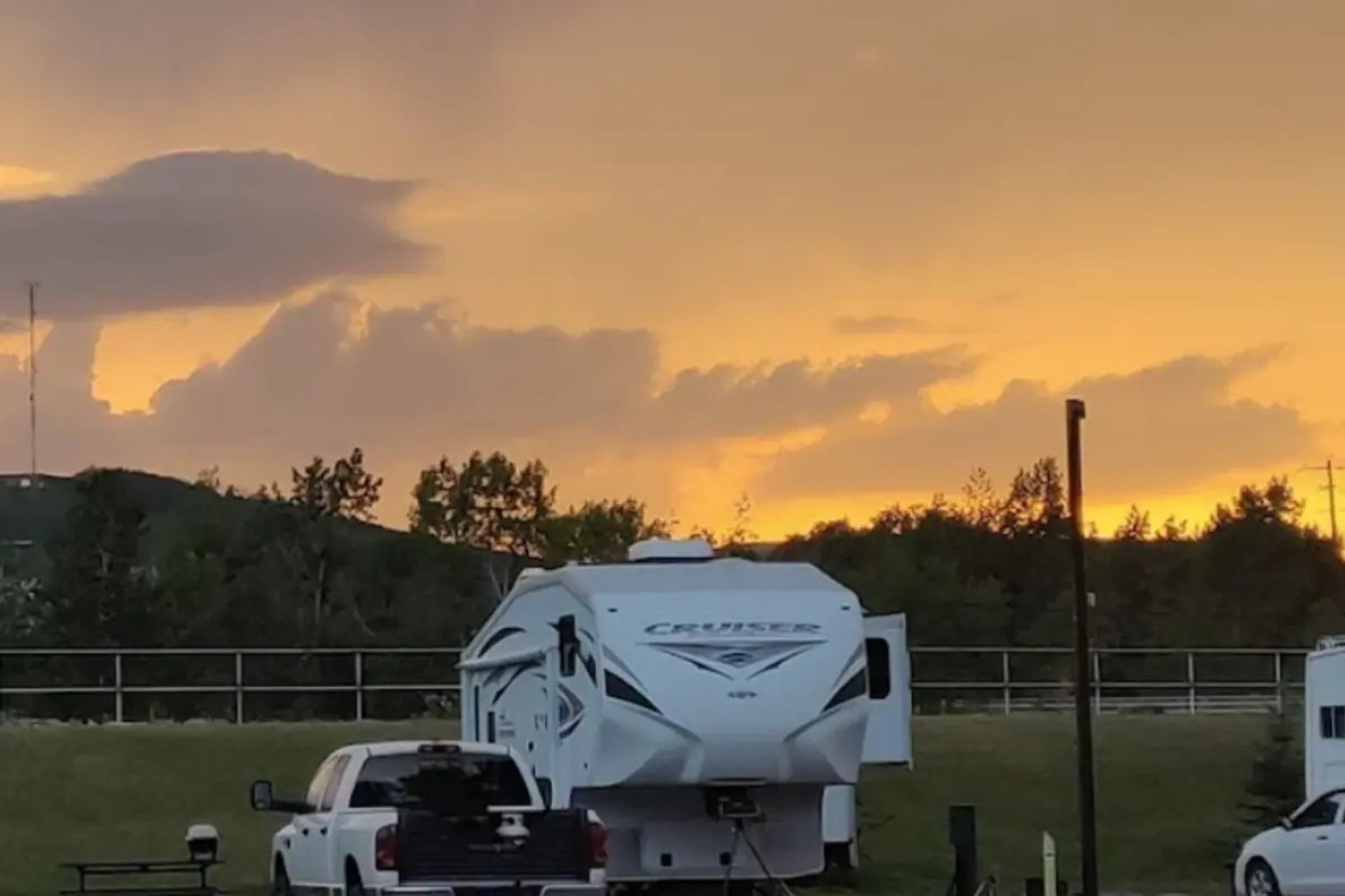 Recreational vehicle and pickup truck parked on grassy campsite under orange sunset sky.