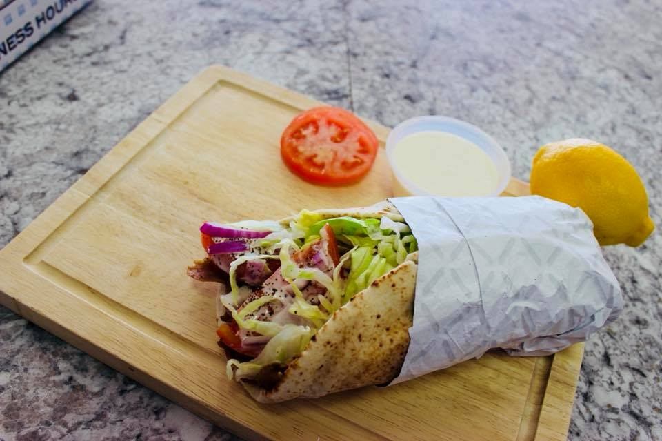 Slice of pizza on a wooden board with lemon, tomato, and dipping sauce cup.