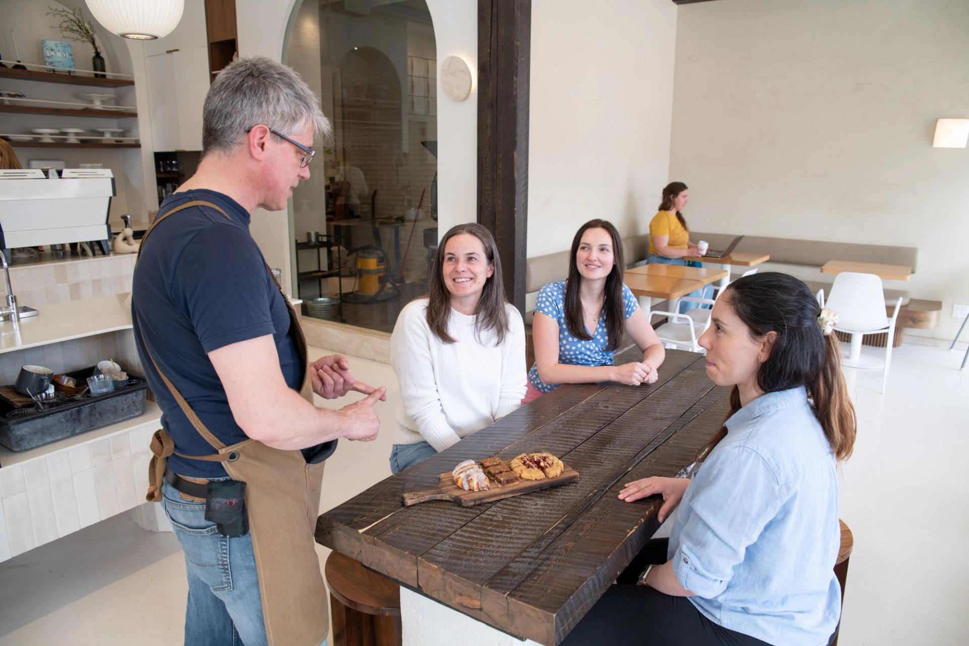 Café server talks with three customers at wooden table with pastries on board.