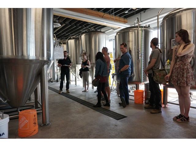 People tour a brewery, standing among large stainless steel fermentation tanks and barrels.