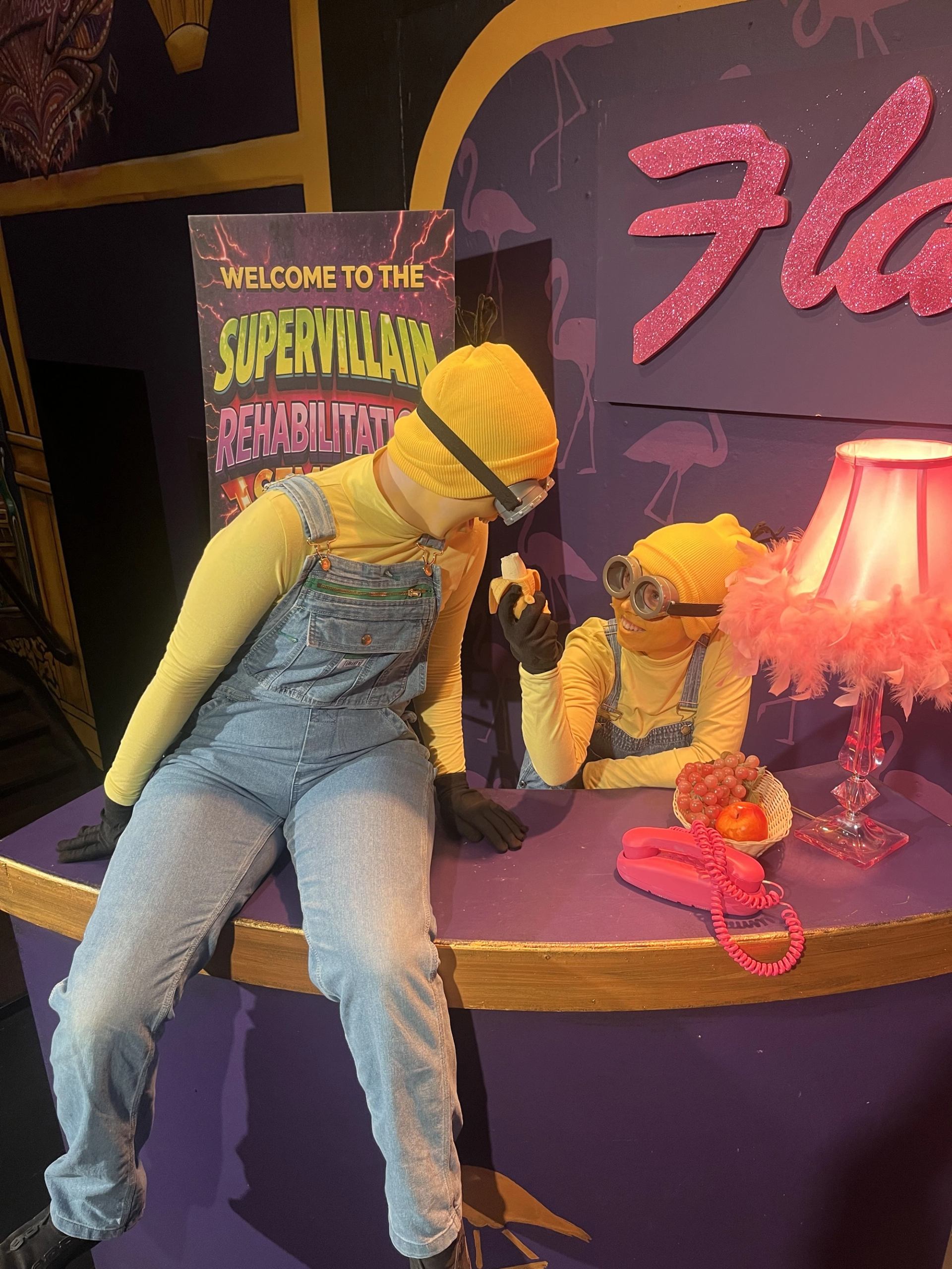 Minion-costumed performers interact beside a pink telephone, fruit basket and feathered lamp.