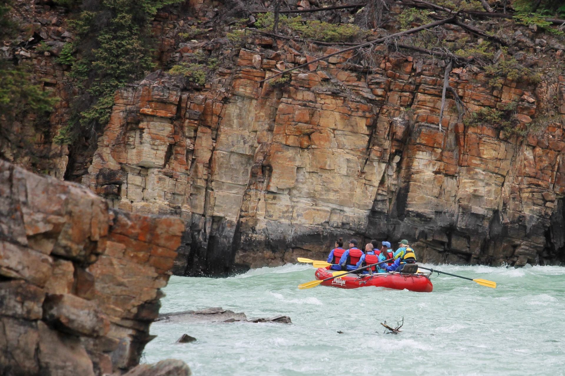 Red inflatable raft with paddlers moves through fast river beside steep rocky cliff.