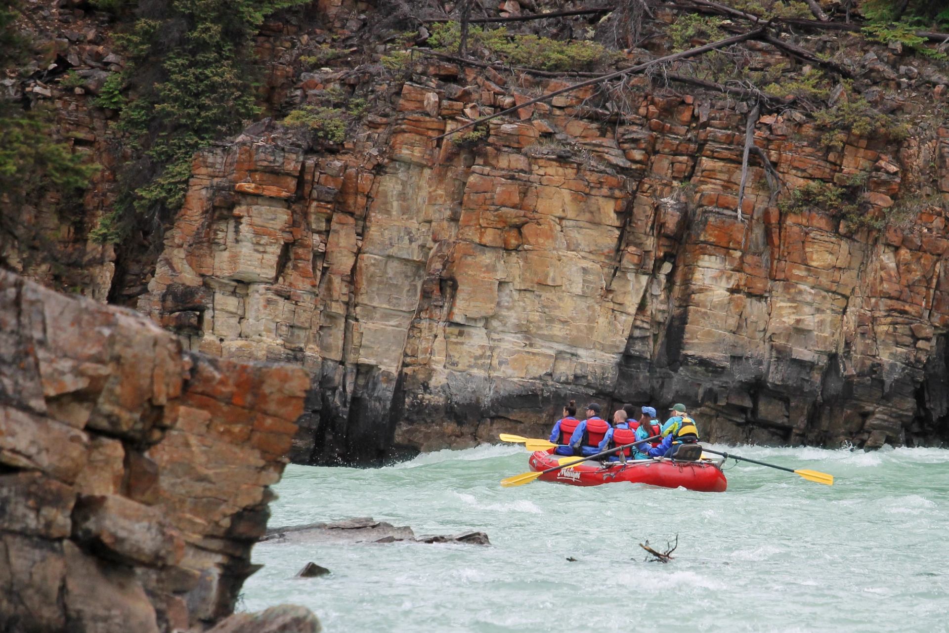 Red inflatable raft with paddlers moves through fast river beside steep rocky cliff.