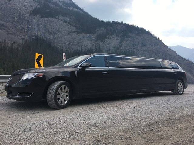 Black Signature Limos vehicle parked on gravel with mountains and curve sign in background.
