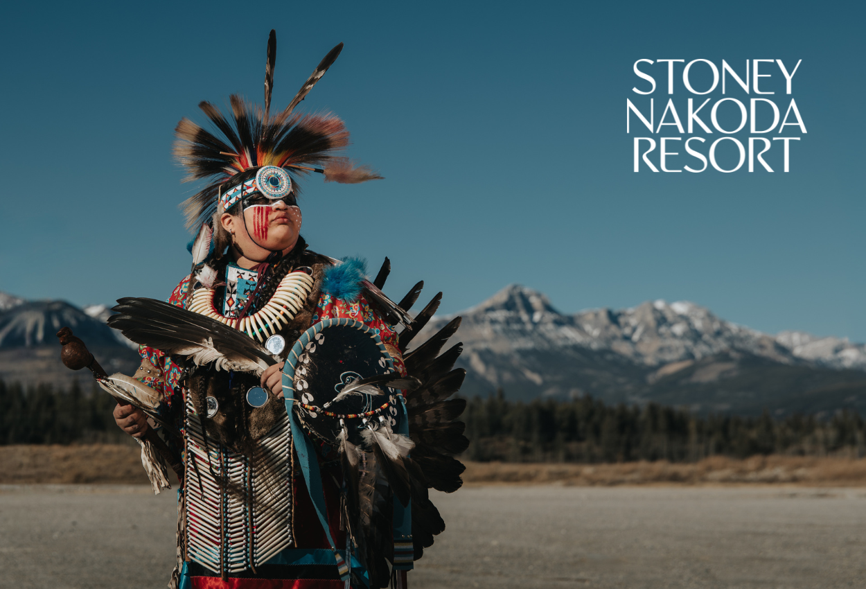 Indigenous dancer in regalia stands outdoors before mountains, with Stoney Nakoda Resort text.