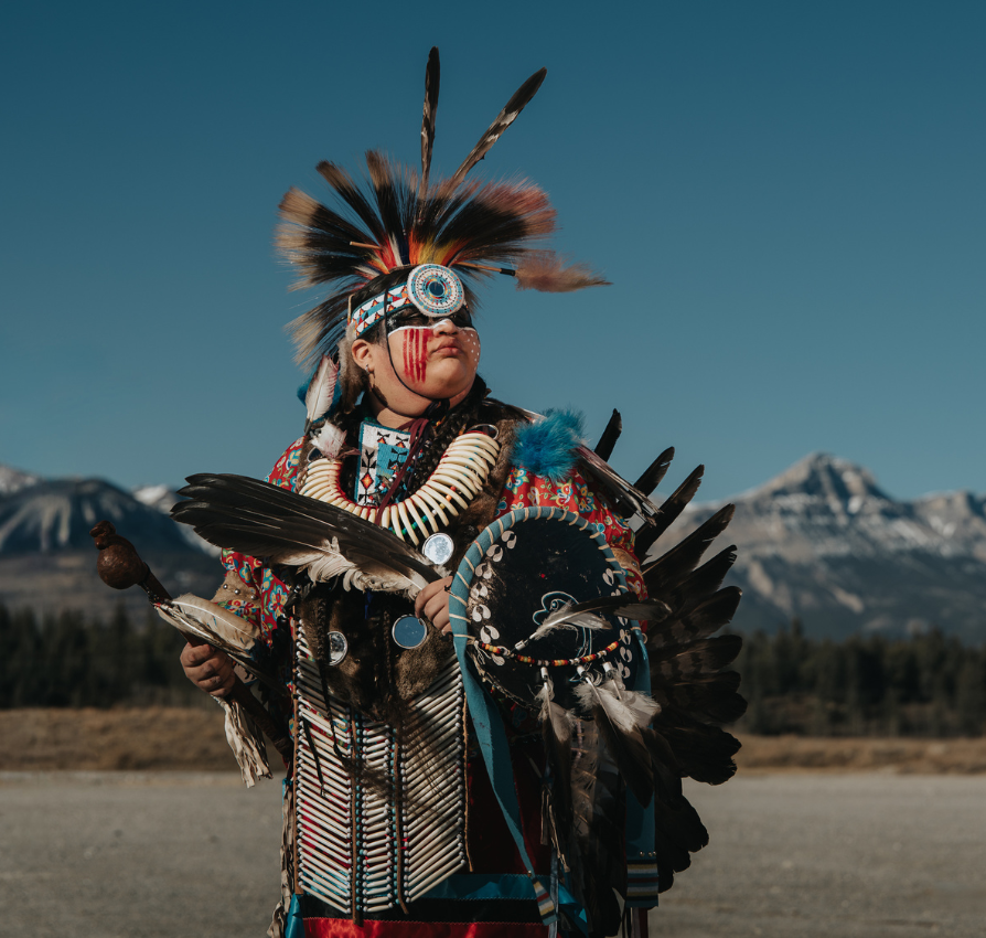 Indigenous dancer in regalia stands outdoors before mountains, with Stoney Nakoda Resort text.