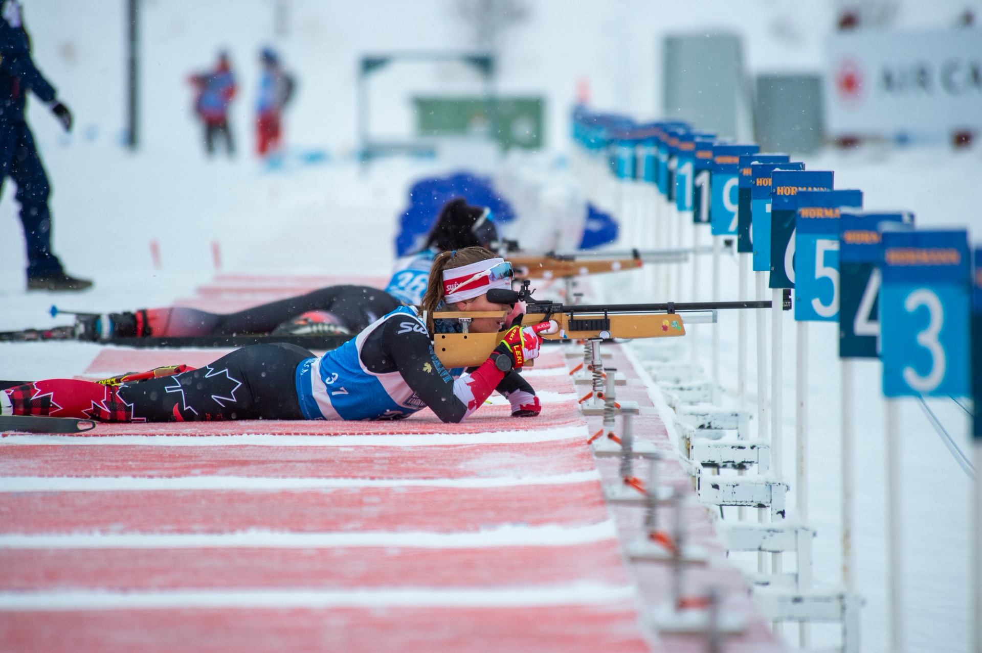 Biathletes lying on snow aiming rifles at targets during a winter competition.