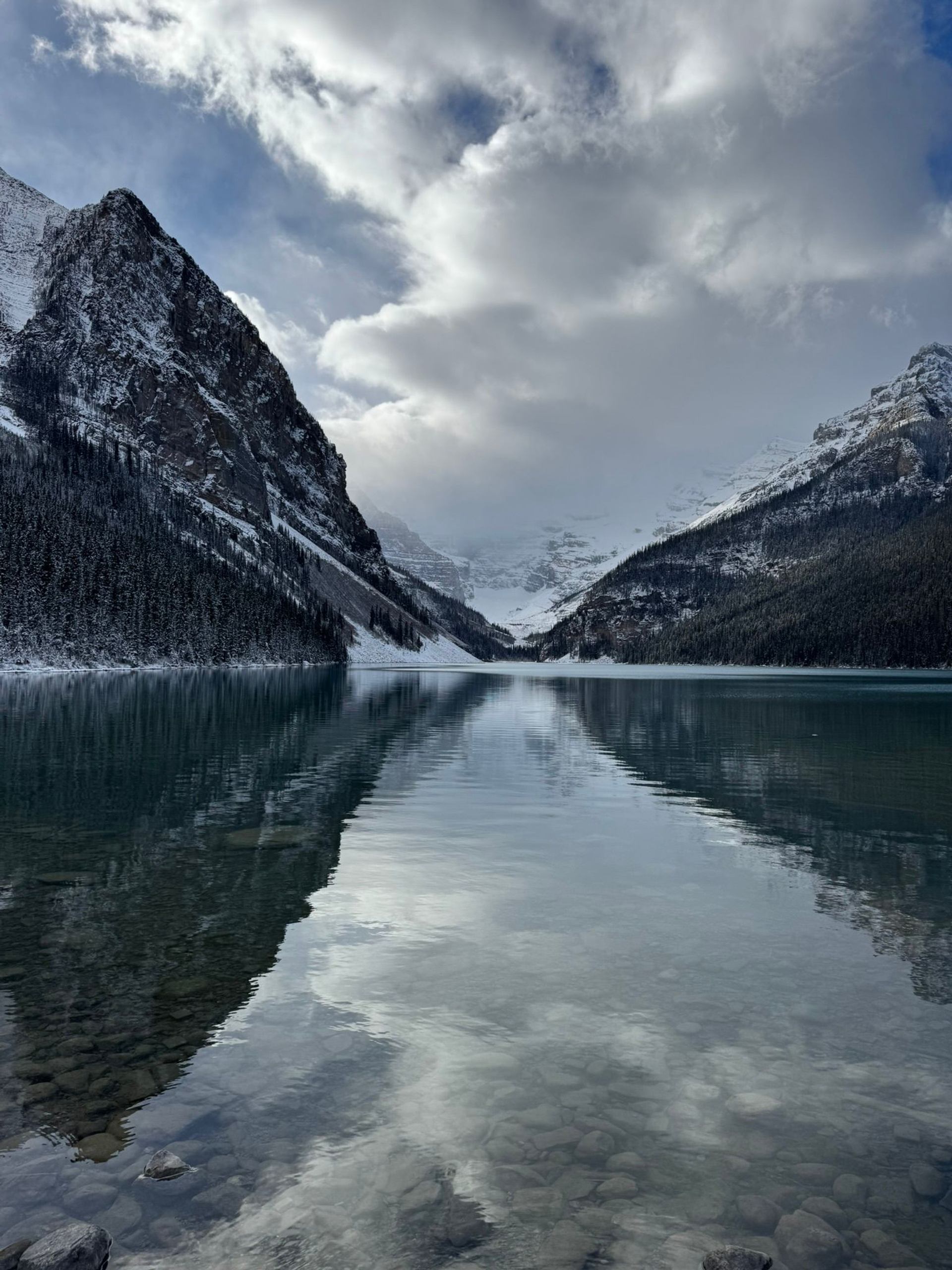 Snowy mountain valley reflected in calm, clear lake with rocky shoreline and clouds.