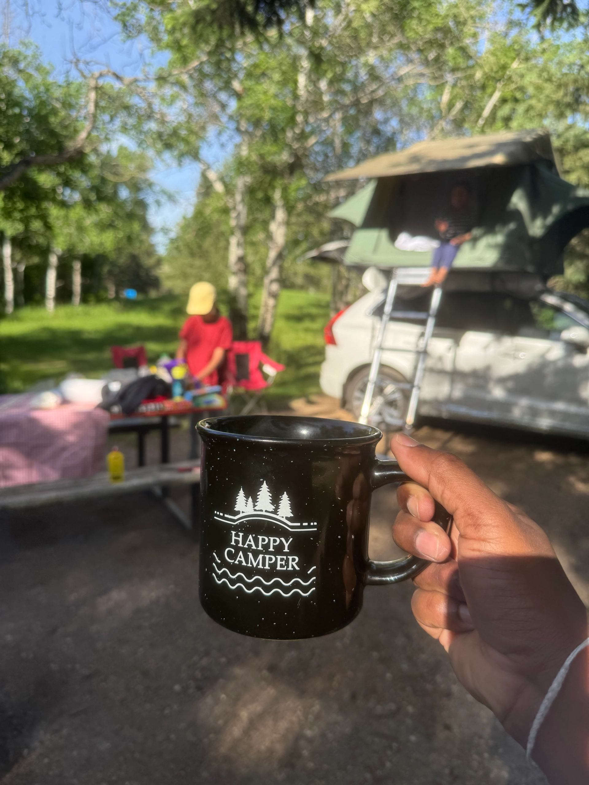 Hand holding Happy Camper mug at campsite with car rooftop tent and trees.