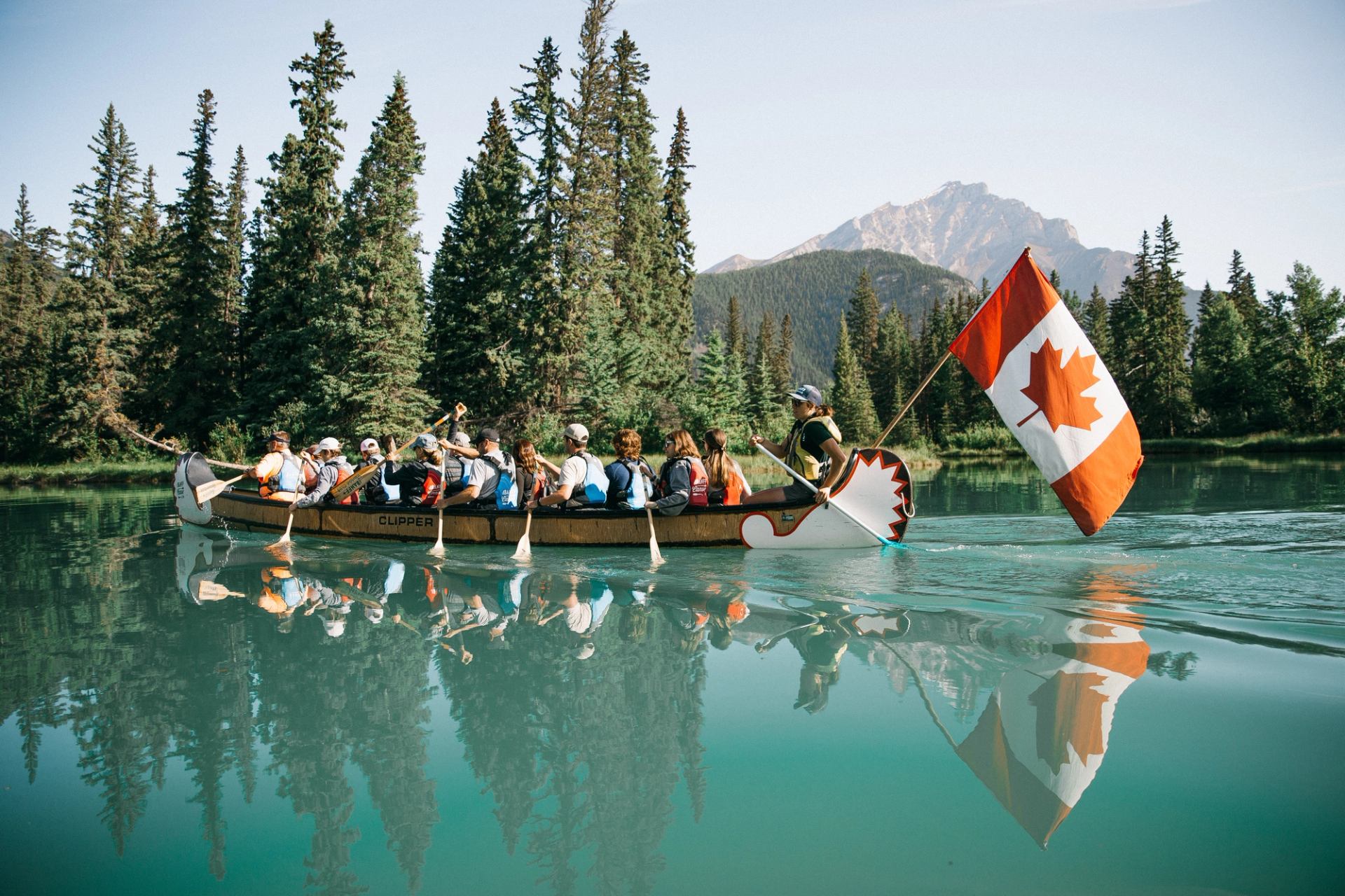 Large canoe of paddlers gliding on clear turquoise water with trees and mountains behind.