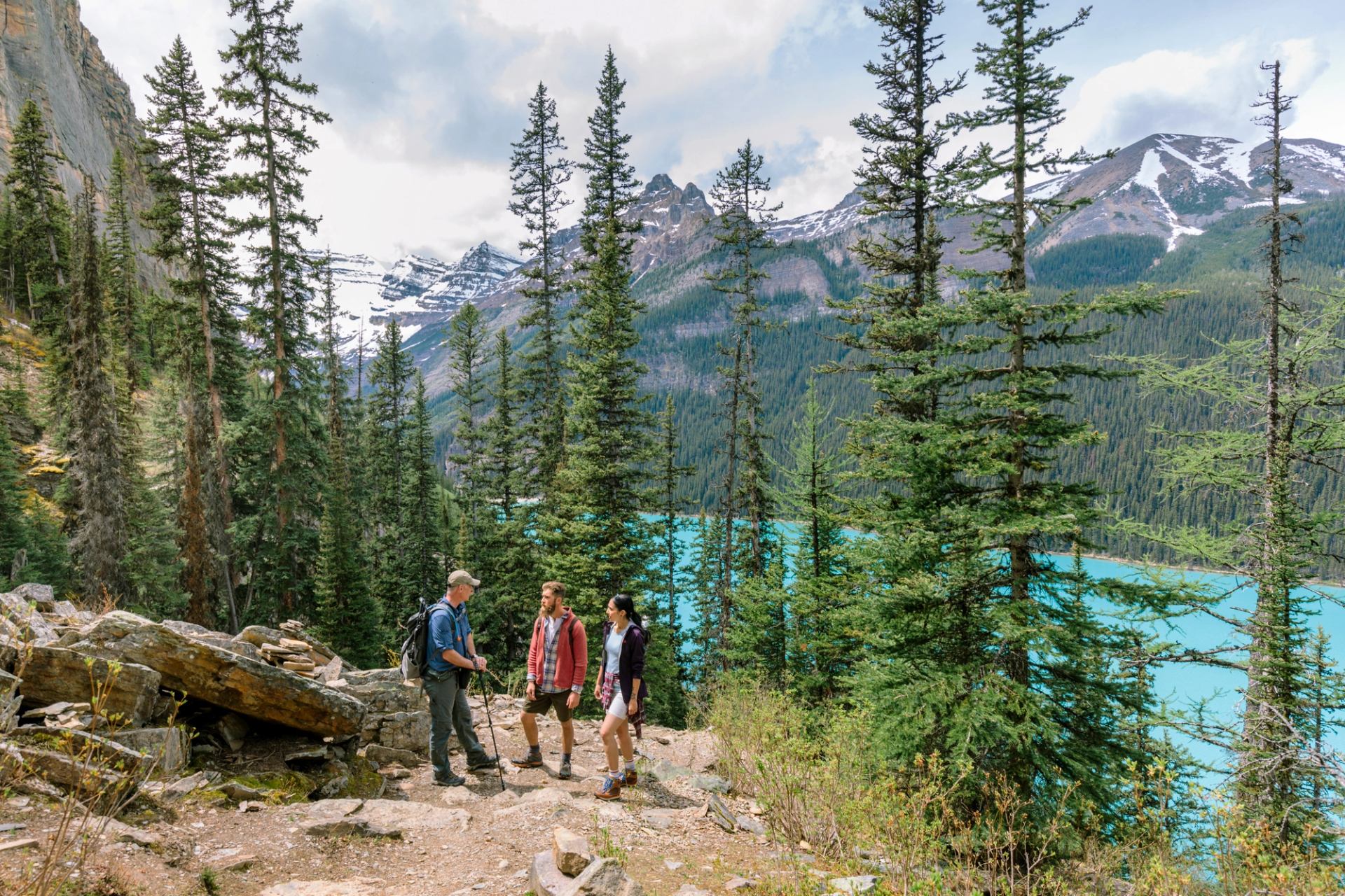 Hikers stand on rocky forest trail overlooking turquoise lake and snow-capped mountain peaks.