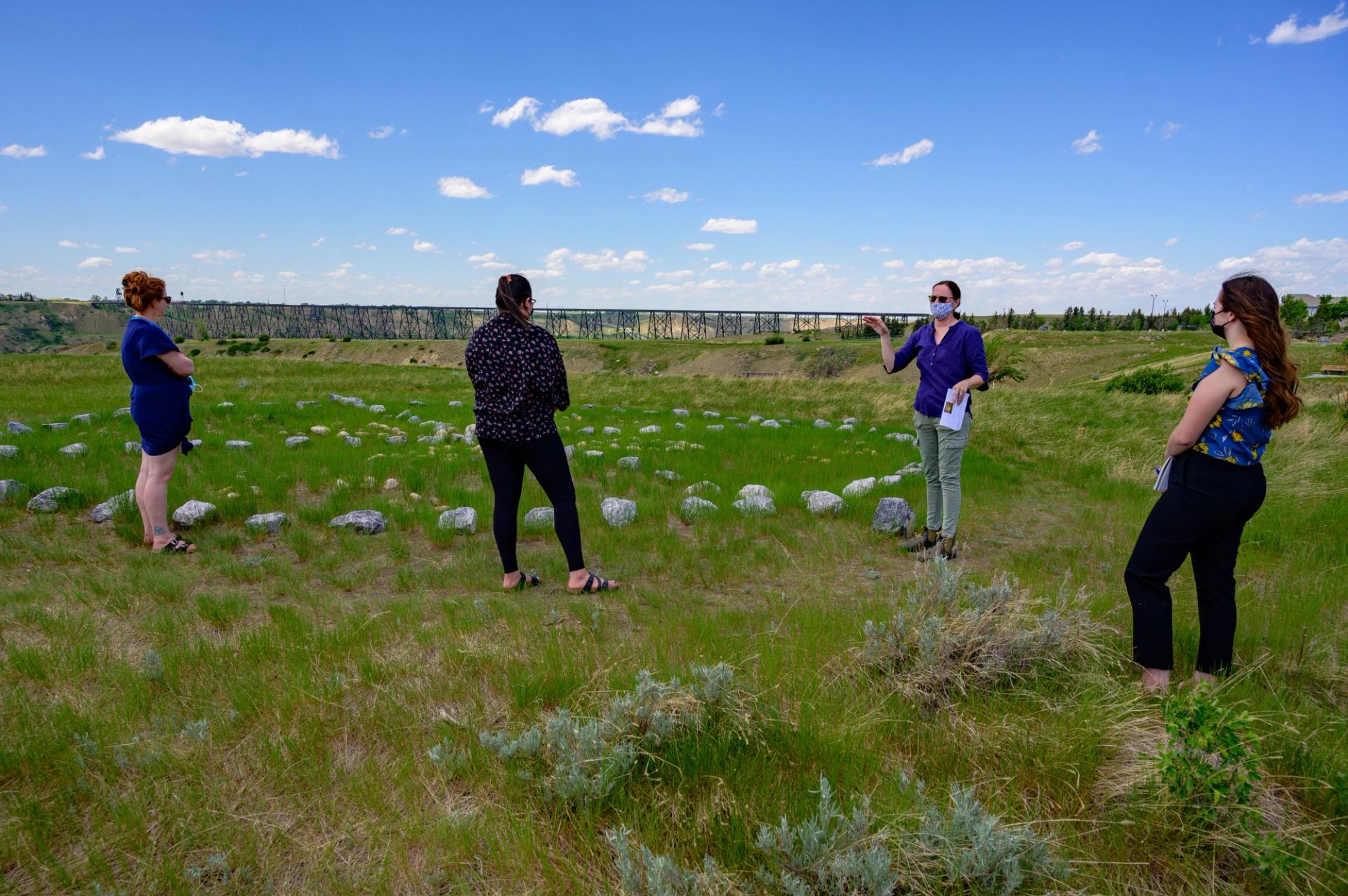 Four people stand distanced in grassy field with stone circle and railway bridge.