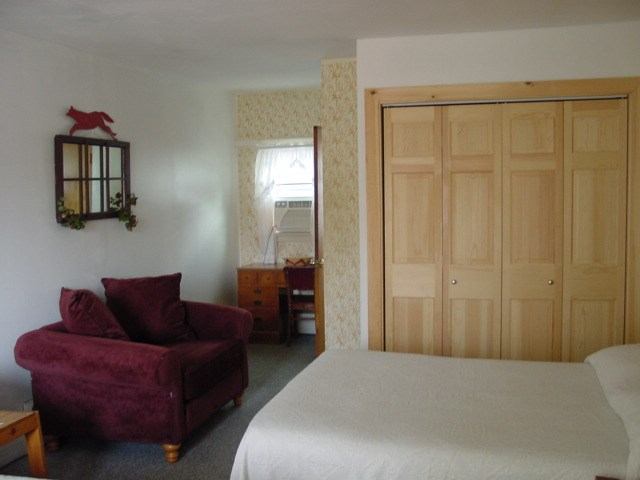 Hotel room with bed, burgundy chaise lounge, wooden closet doors and desk by window.