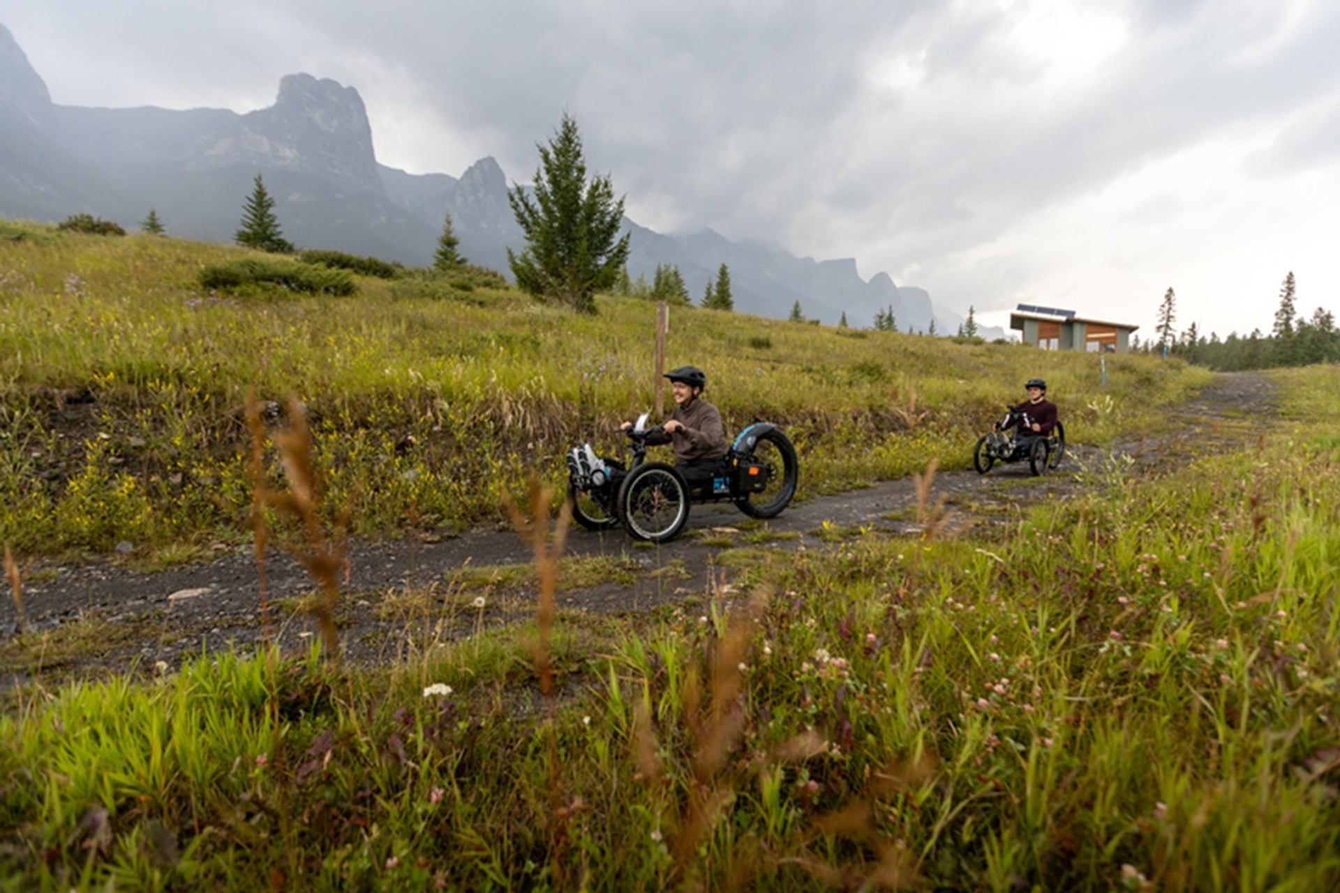 Two people ride adaptive off-road trikes along a grassy trail with distant mountains.
