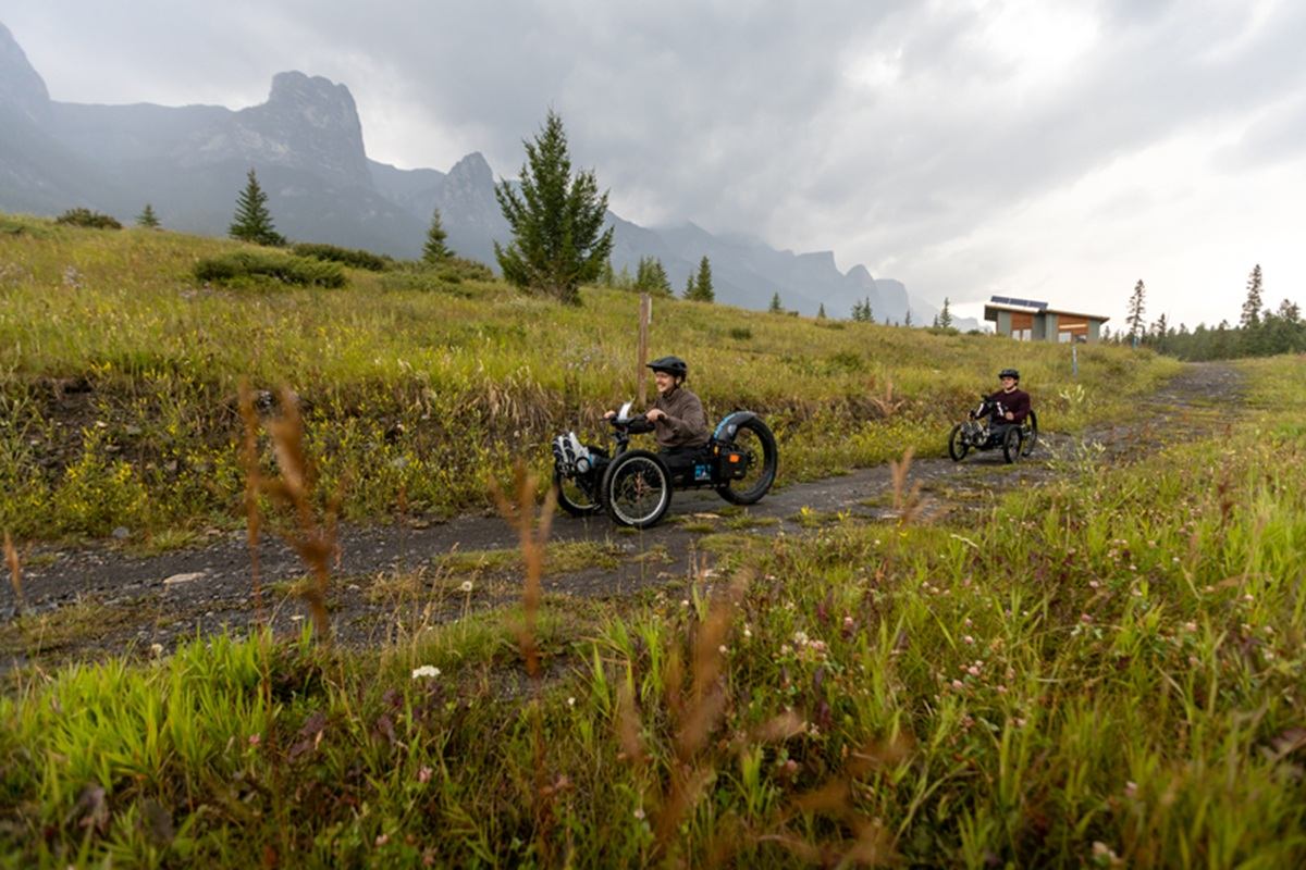 Two people ride adaptive off-road trikes along a grassy trail with distant mountains.